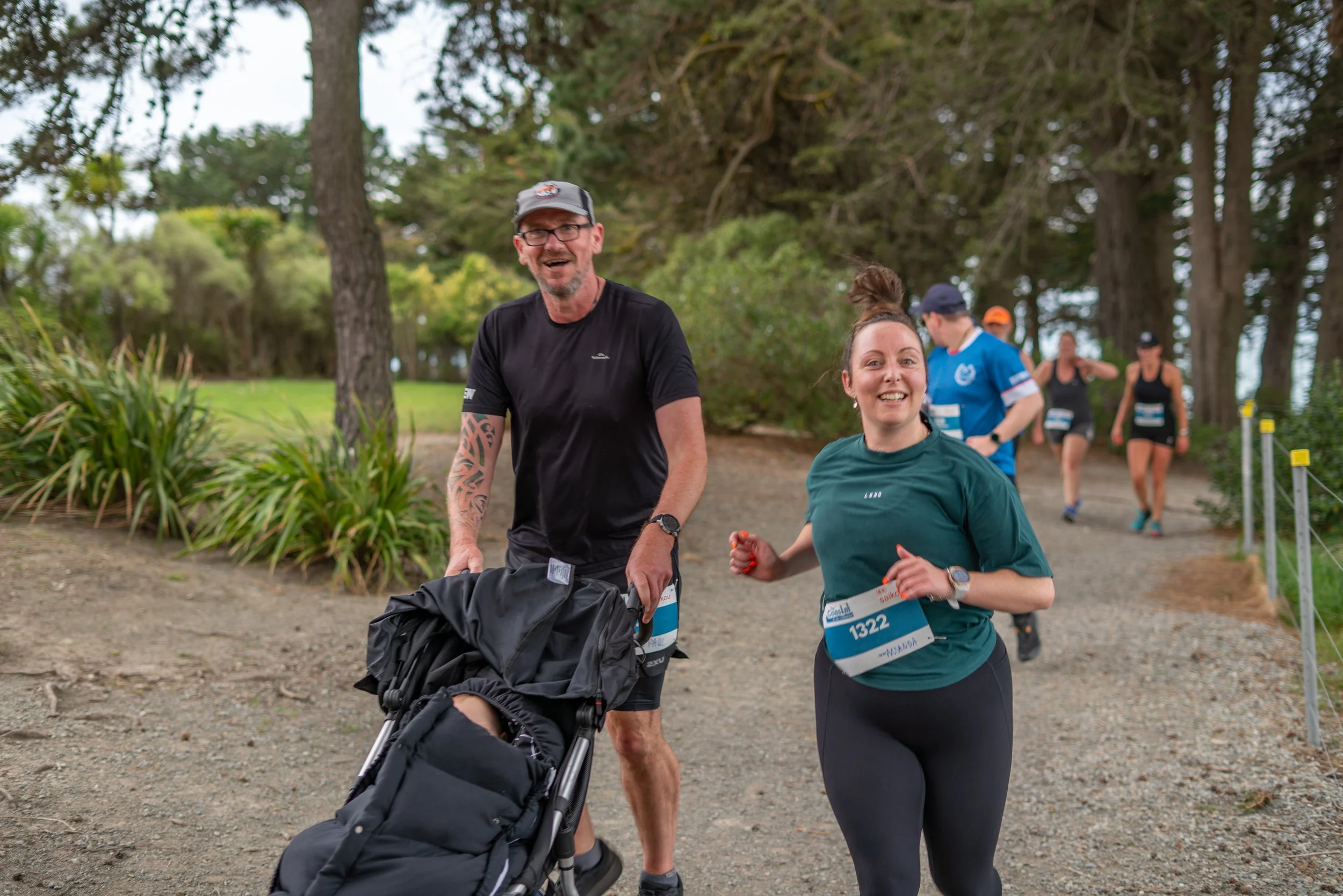 People participating in a race or running event in a park, with one woman in a green shirt and a man pushing a stroller, smiling as they run on a dirt path surrounded by trees and greenery.