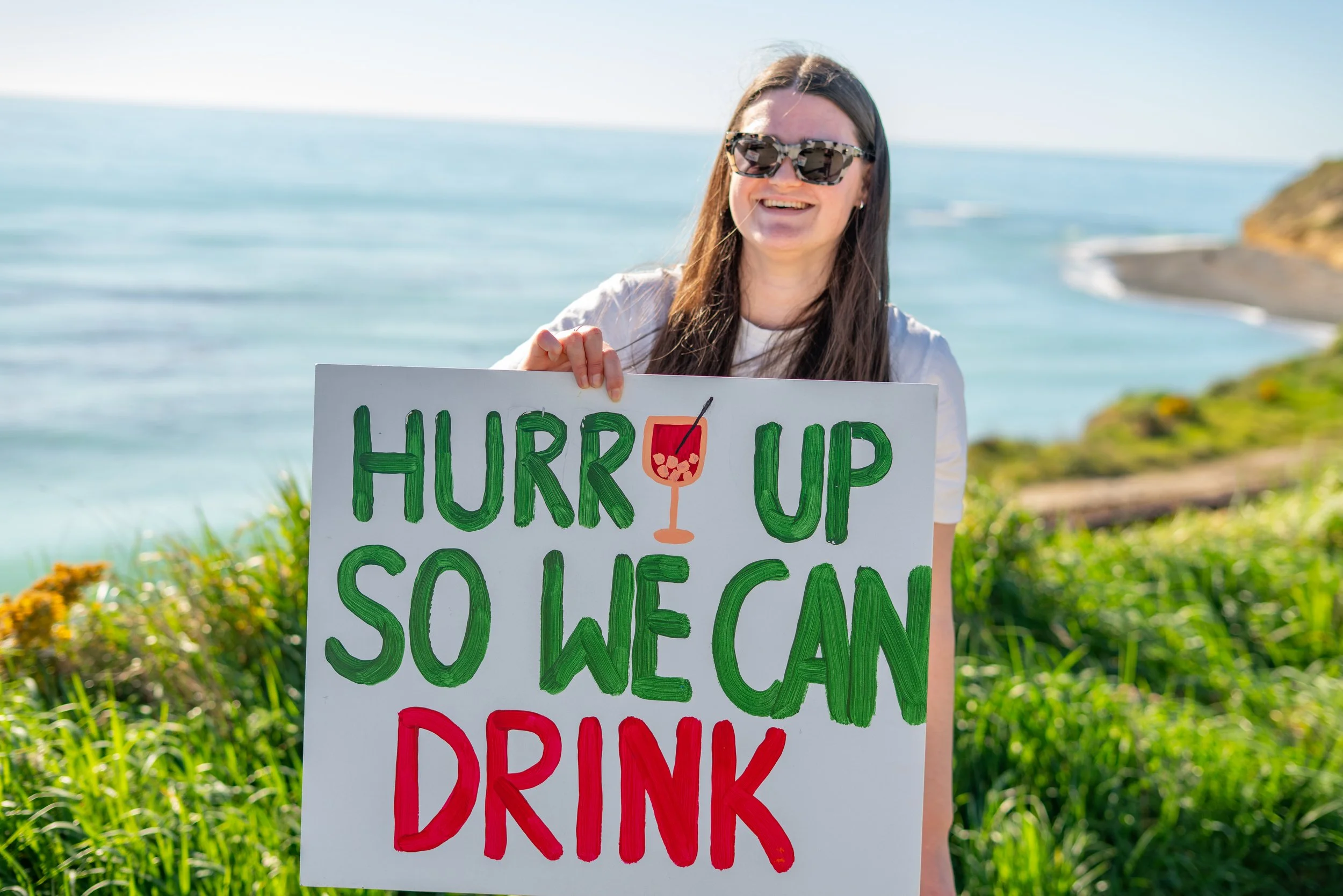A smiling woman wearing sunglasses holding a colorful sign that says 'HURR UP SO WE CAN DRINK', with a drawing of a cocktail glass, at a beach or coastal area.
