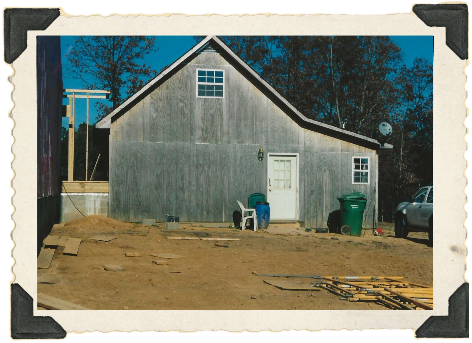 The First Cabin: A picture of the first phase of the current Hollowell home. It is made of untreated wood. There are trees behind the house and trash cans, a chair and construction equipment in front of it.