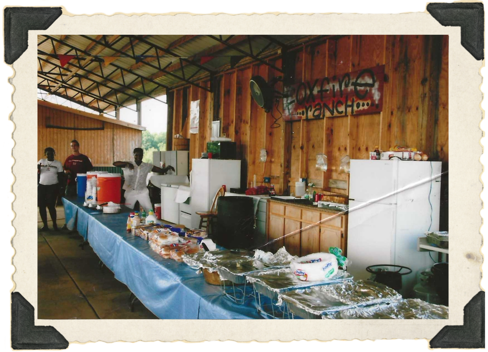 A long table is set wth blue table cloths, chafing dishes, blue and orange plastic drink dispensers. There is a wood paneled wall to the right with a red and with sign that says “Foxfire Ranch” in Black lettering.