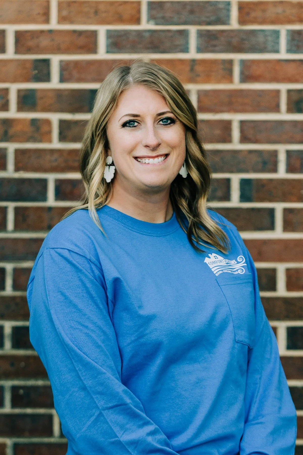 A woman with blonde hair, wearing a blue collared shirt and hoop earrings, standing against a brick wall and smiling.