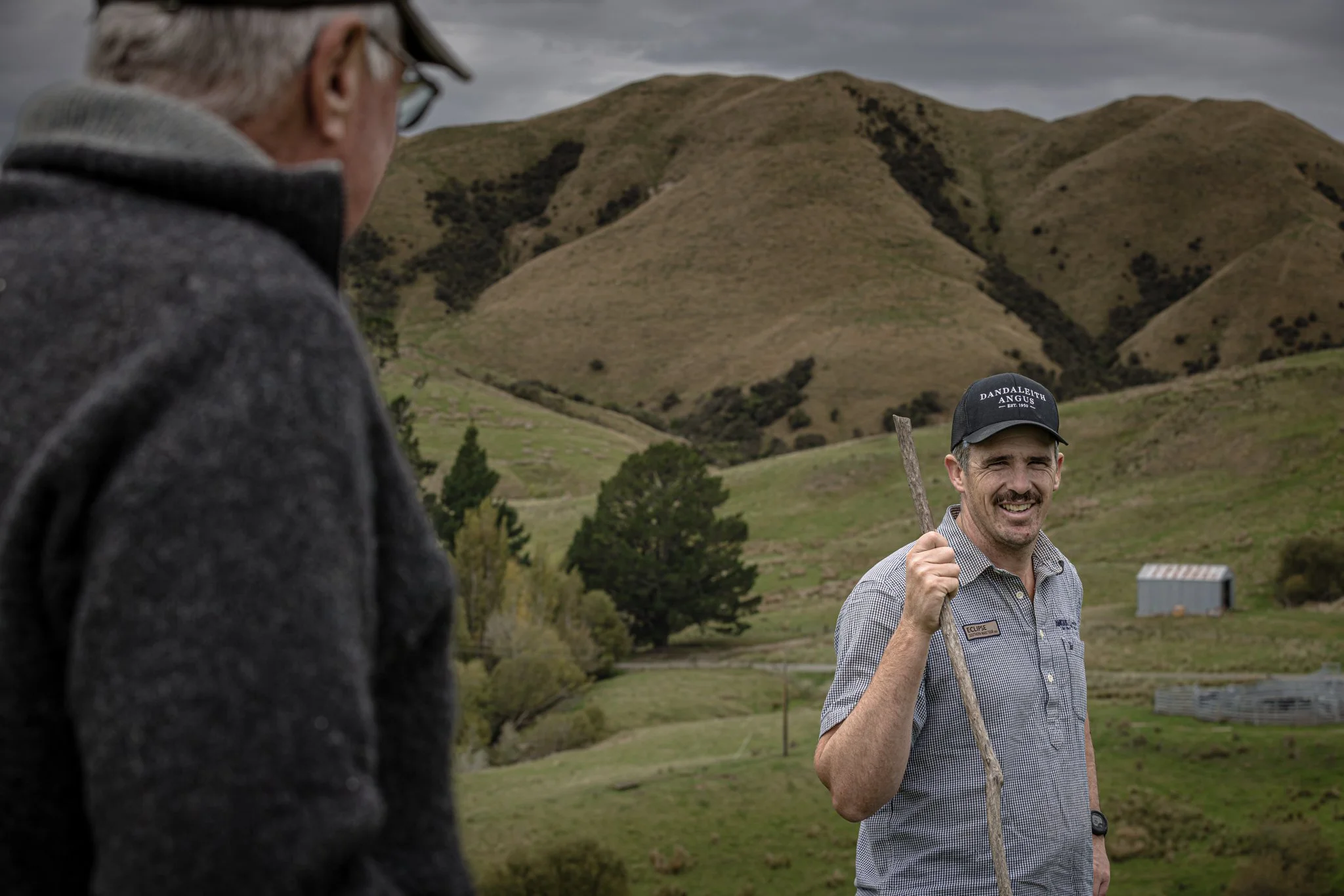A man smiling and holding a stick outdoors in a grassy, hilly landscape with trees and a small building in the background. Another person is partially visible in the foreground.