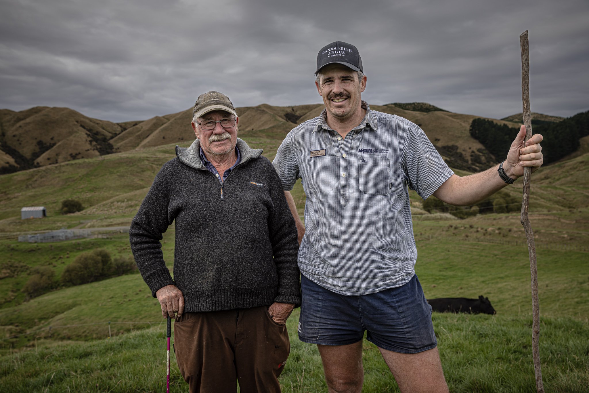 Two men standing outdoors on a hilly landscape under cloudy sky, one with a walking stick and the other with a golf club.
