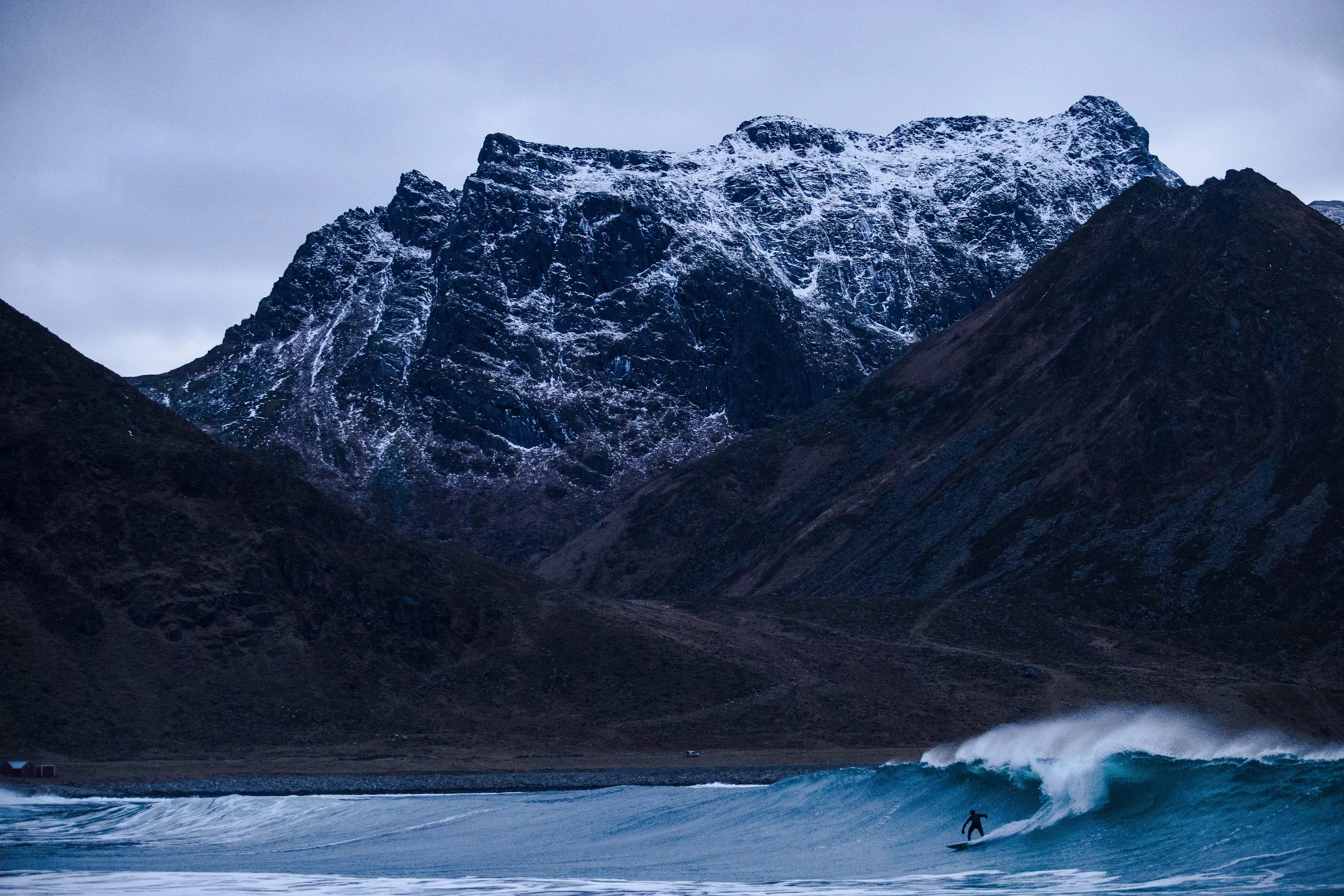 Une scène outdoor en Norvège avec un surfeur près des vagues, face à une montagne enneigée et sombre sous un ciel nuageux dans la tempête