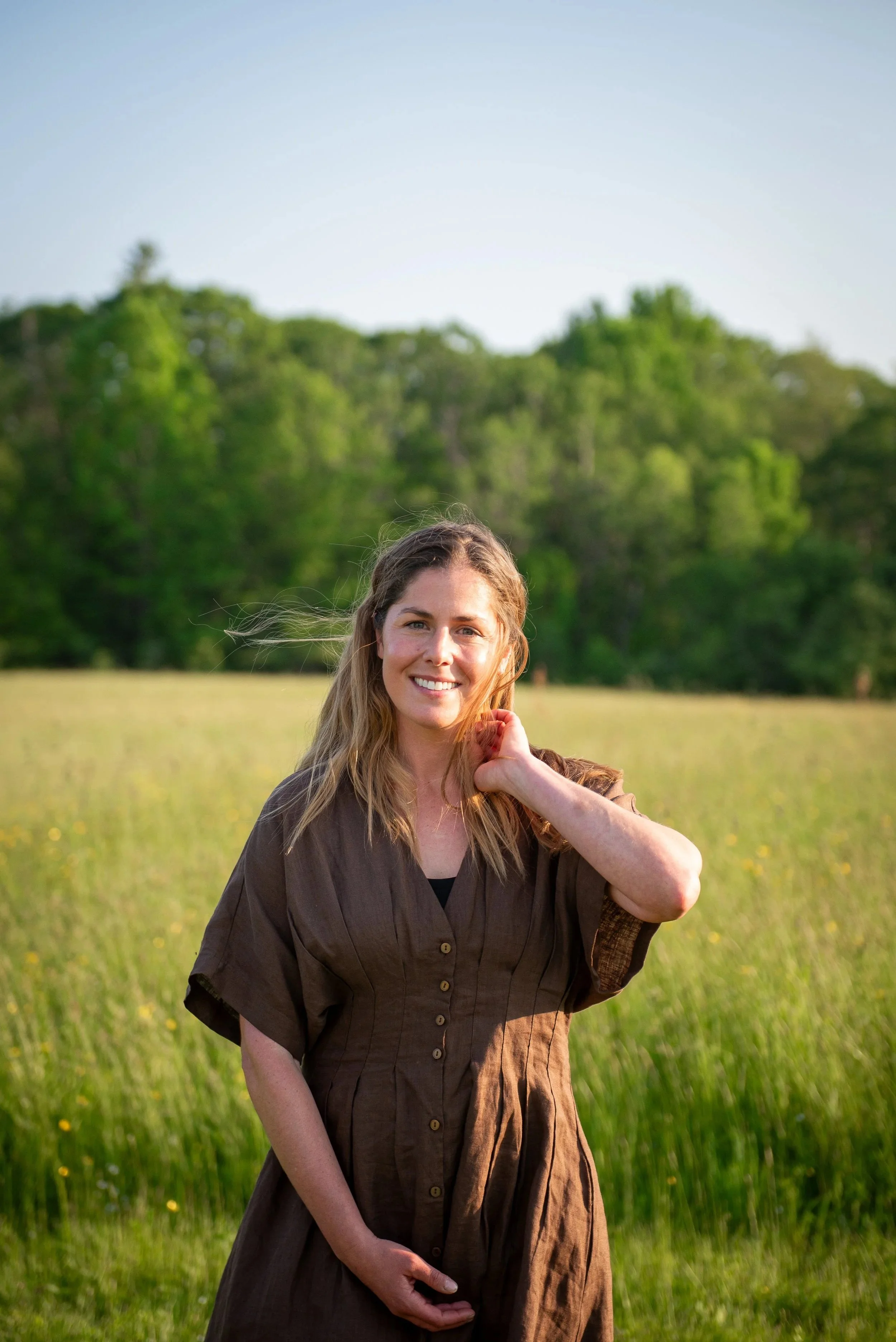 A woman with long blonde hair smiling while standing in a green field with trees in the background on a sunny day.