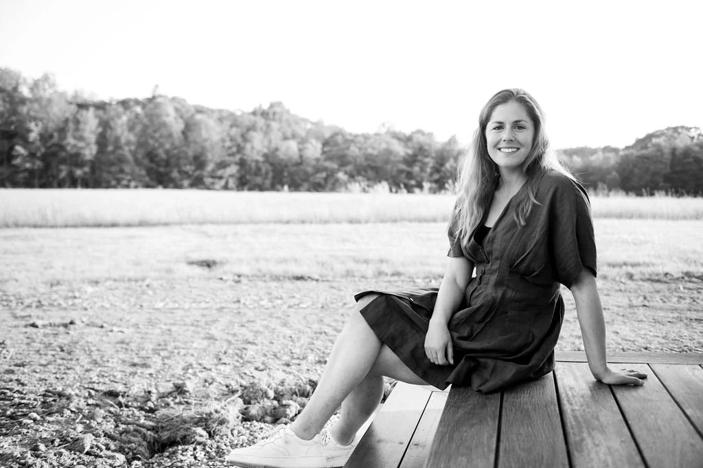 A woman sitting on a wooden dock outdoors, smiling at the camera with a scenic natural background.