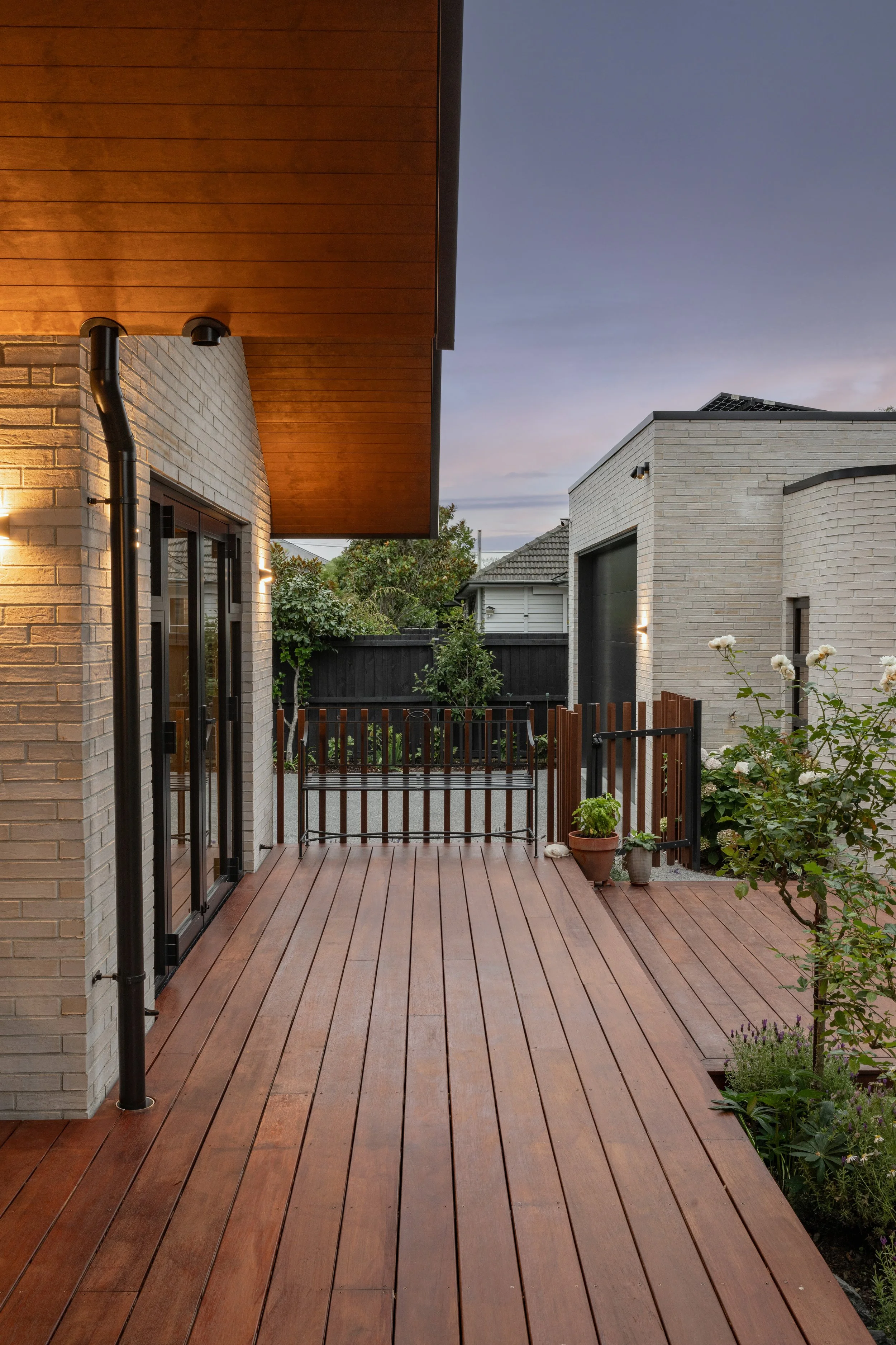 View of a wooden outdoor deck with potted plants, a garden bench, and a modern house with a light-colored brick exterior. The house has black sliding glass doors, and there is a garden with greenery and flowers visible in the background. The sky is p