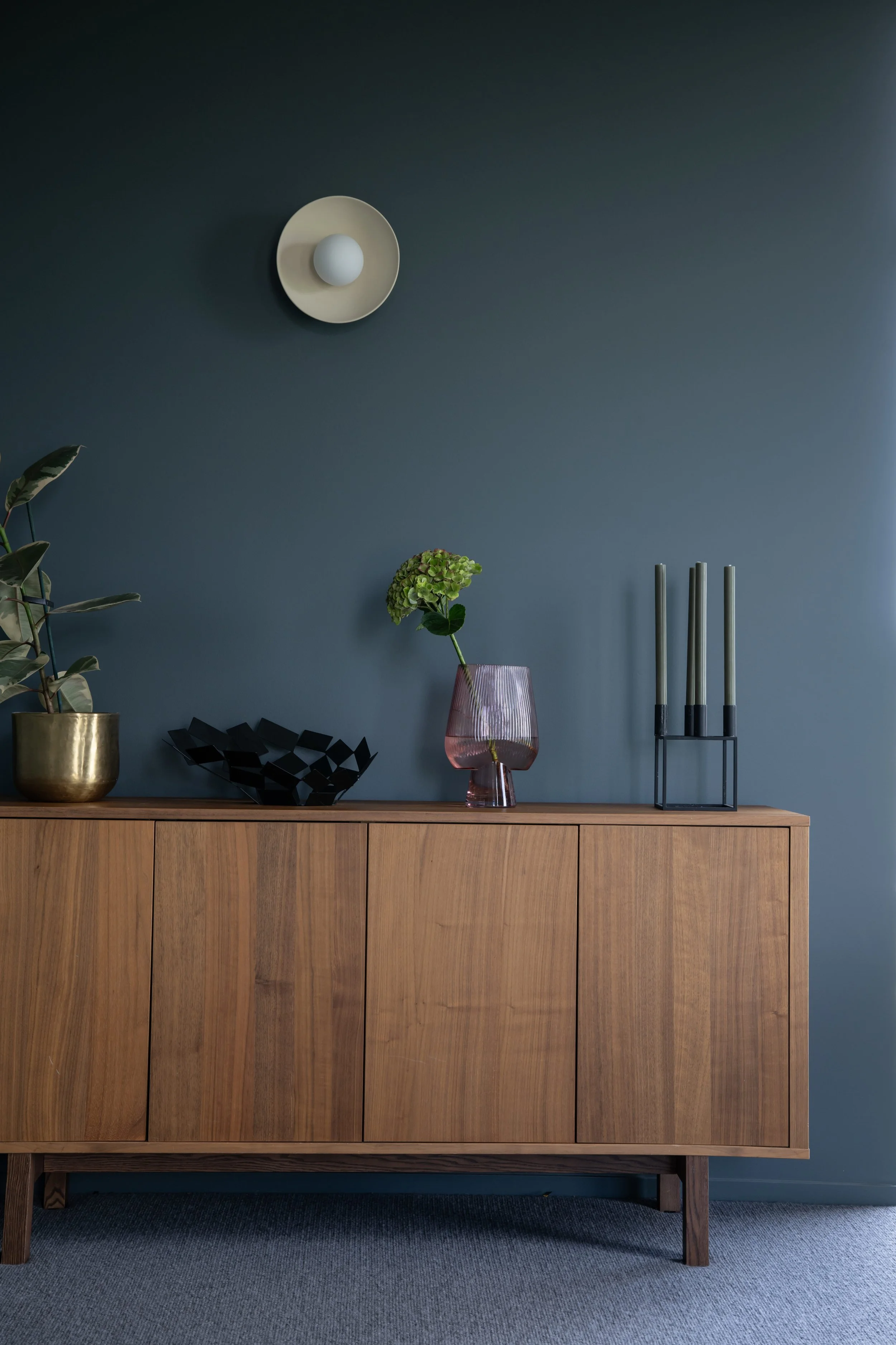 A wooden sideboard with a dark blue wall behind it. On the sideboard there are several decorative items, including a gold pot with a large green plant, a black geometric sculpture, a purple vase with a green flower, and a black metal stand holding th