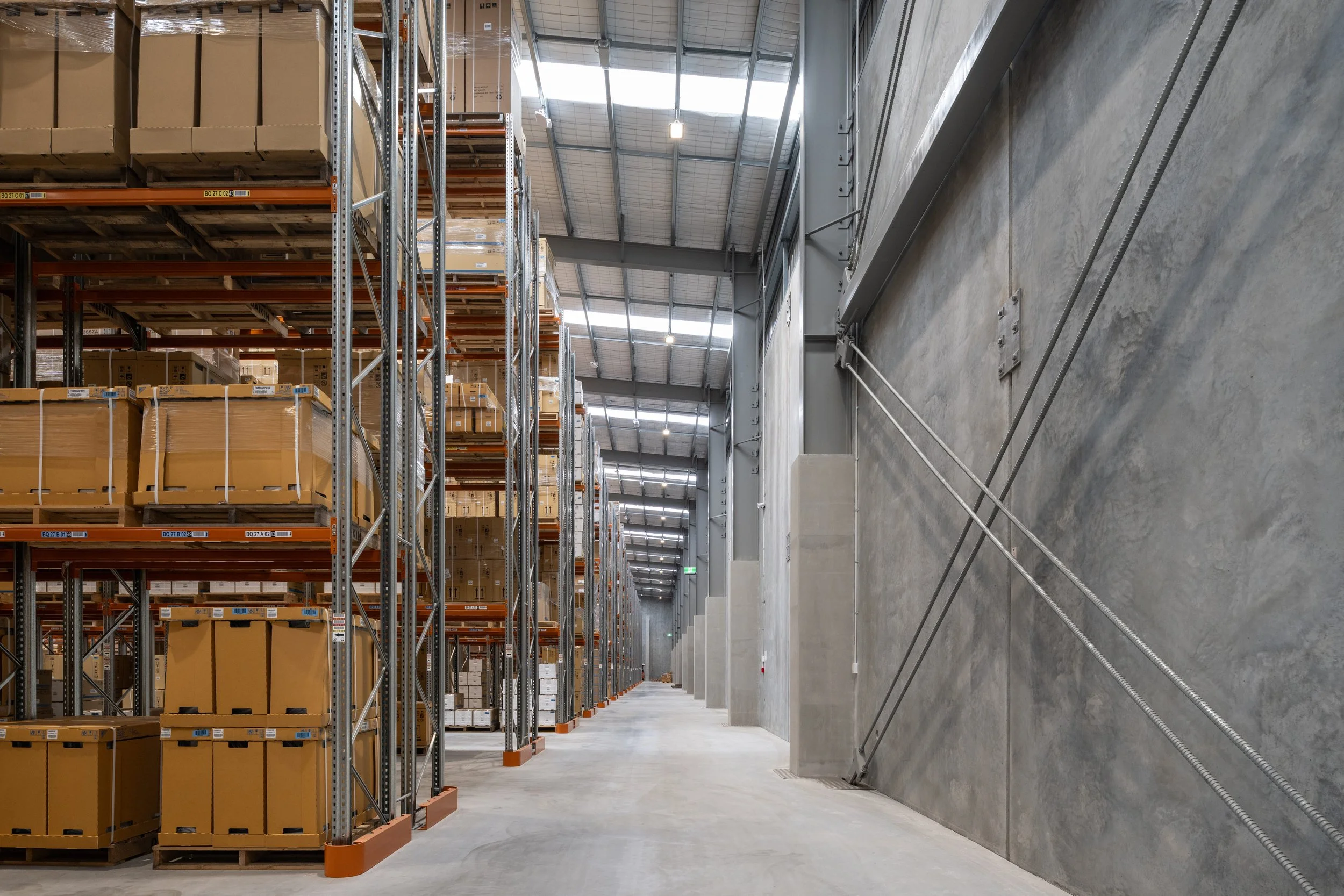 Warehouse aisle with metal shelving filled with cardboard boxes and pallets, concrete walls, high ceiling with skylights, and industrial lighting.