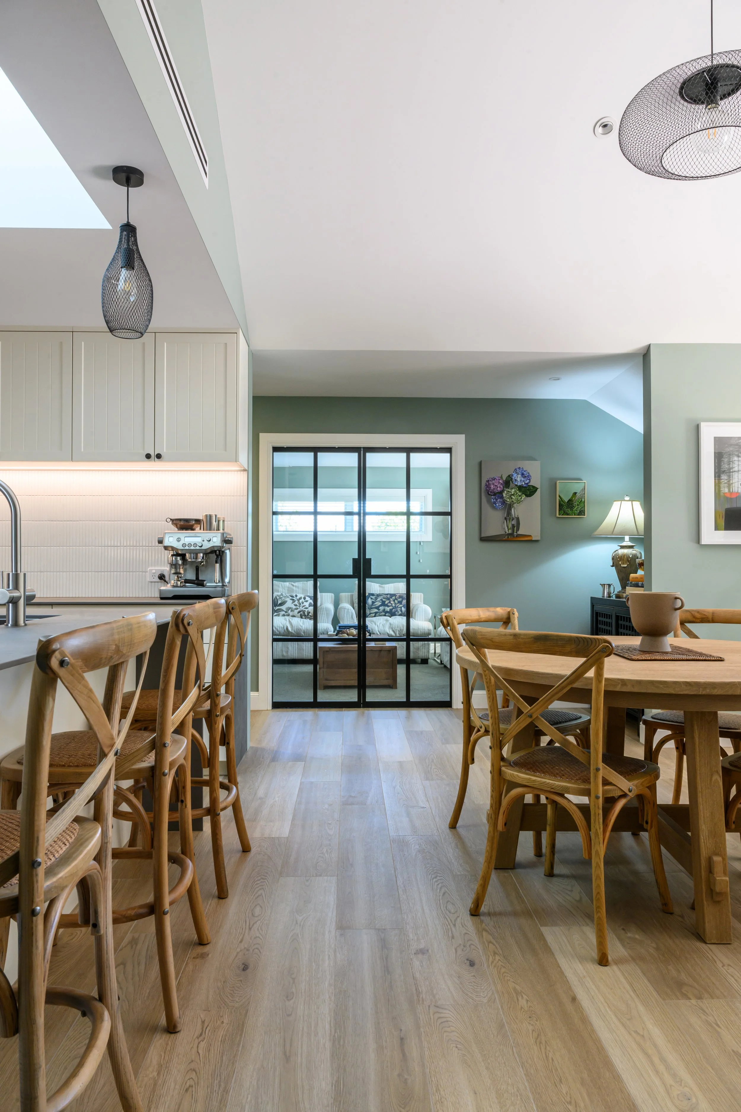 Open-concept kitchen and dining area with wooden chairs, pendant lights, and a glass sliding door leading to a living room with a sofa and wall art.