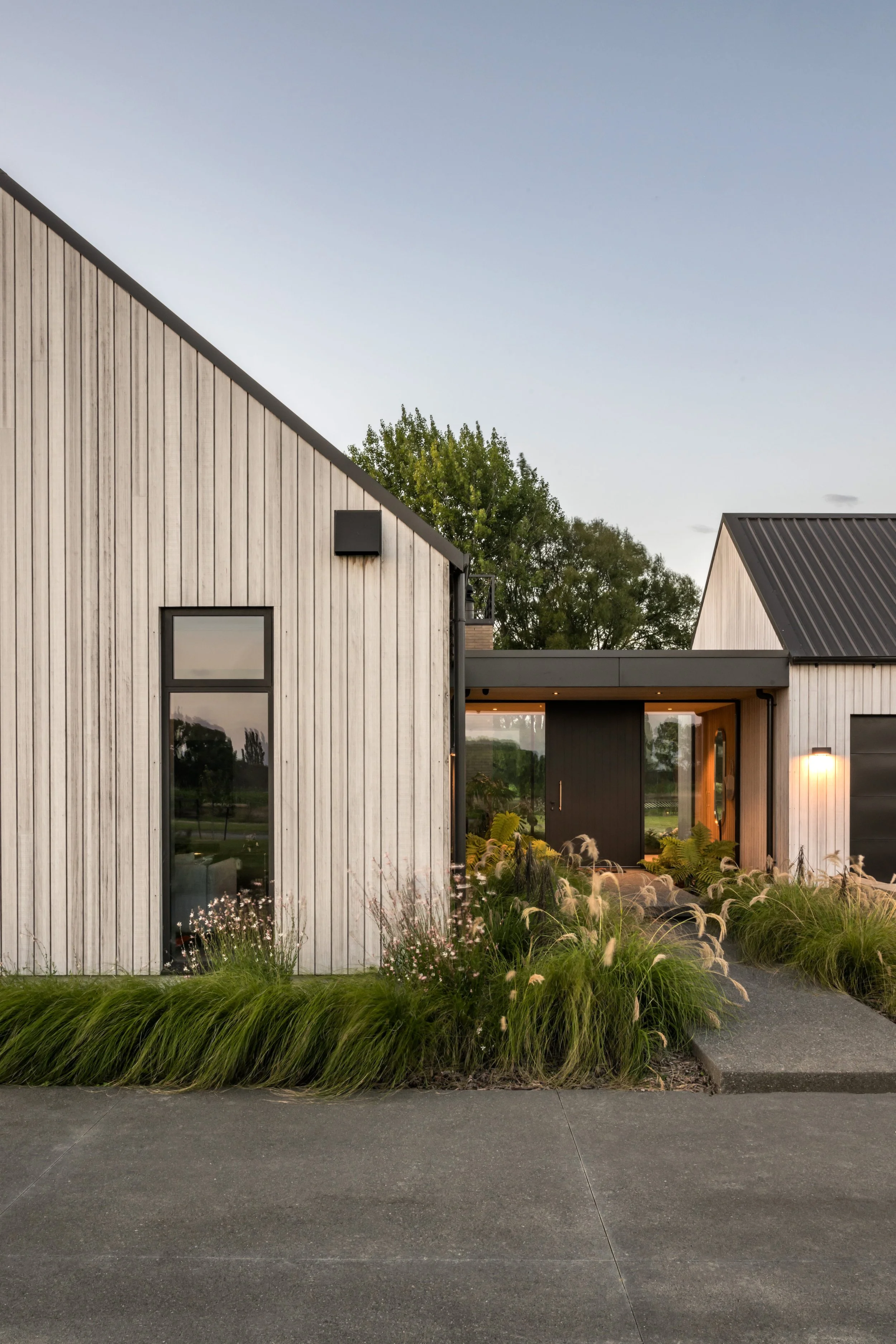Modern house with wooden exterior, sloped roof, large window, and landscaped garden with grass and plants at dusk.