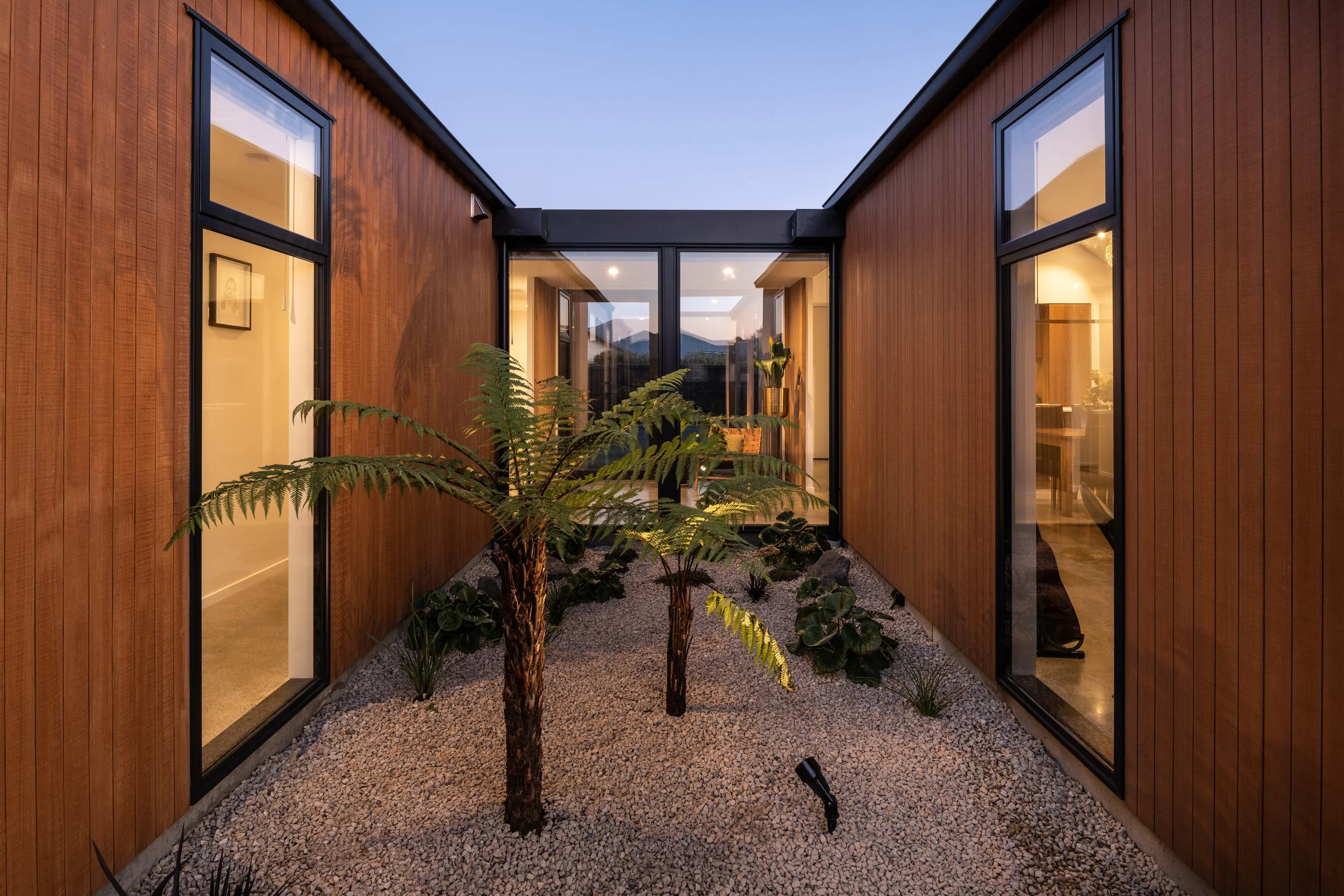 Interior courtyard of a modern house with wooden walls, large glass windows, a small gravel ground, and indoor plants, with interior lights on inside the house.
