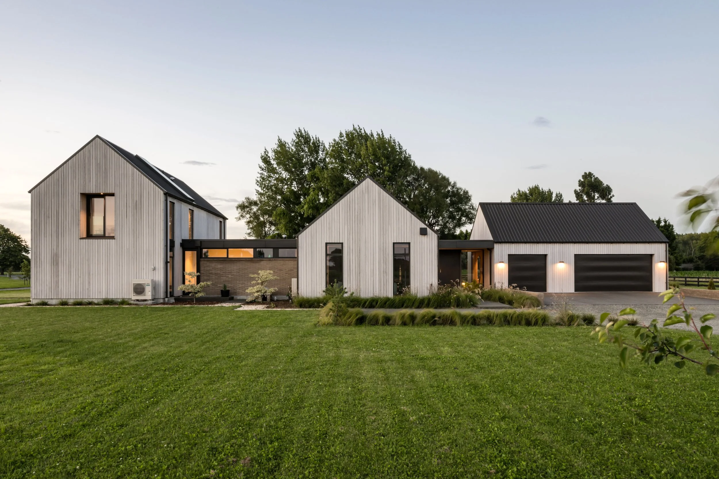 Modern house with white vertical siding, large windows, and a dark metal roof, surrounded by a well-maintained green lawn and trees, during dusk with outdoor lights on.