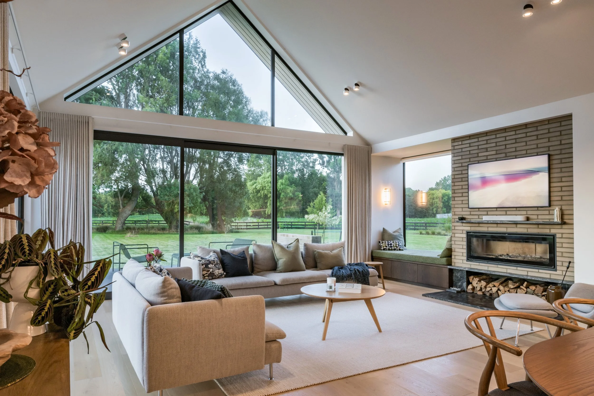 Modern living room with large windows showing green outdoor scenery, featuring a beige sofa with decorative pillows, a round wooden coffee table, and a built-in fireplace with a TV above it. There are several indoor plants and a dining area with a wo