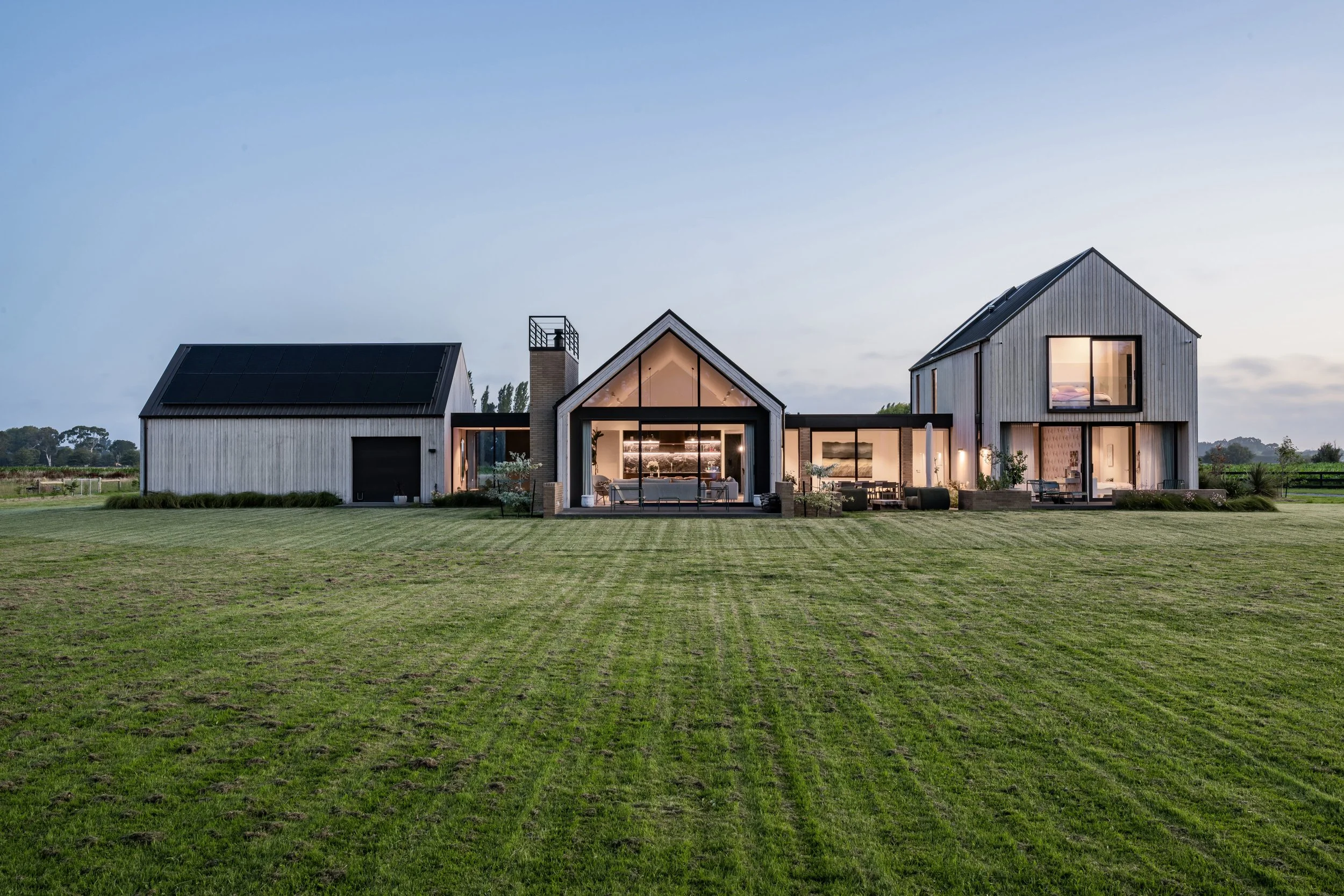 A modern farmhouse with a large glass front, wooden siding, and a metal roof, set in a spacious grassy yard at dusk with interior lighting visible.