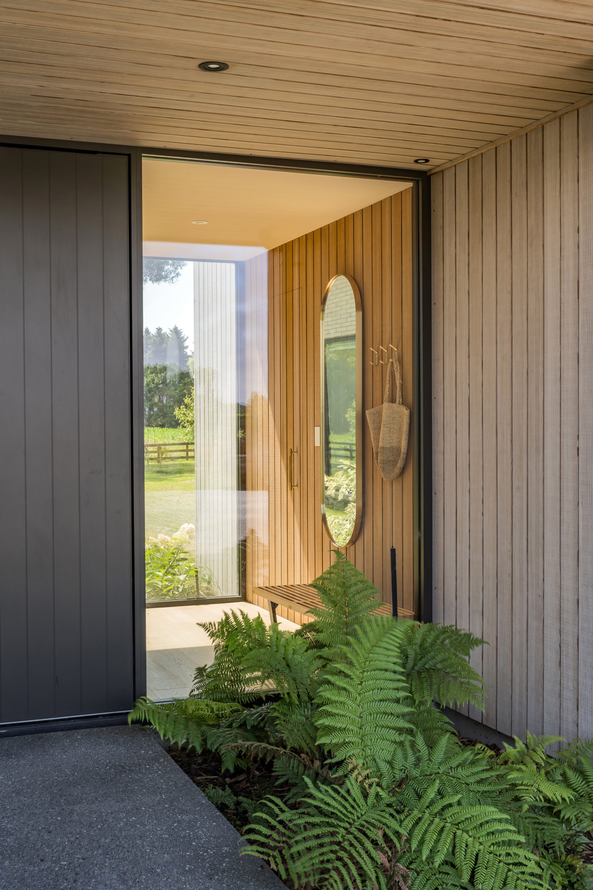 View of a modern entrance with wood paneling, a large oval mirror, a small wooden bench, hanging bag, and lush green ferns outside through glass door.
