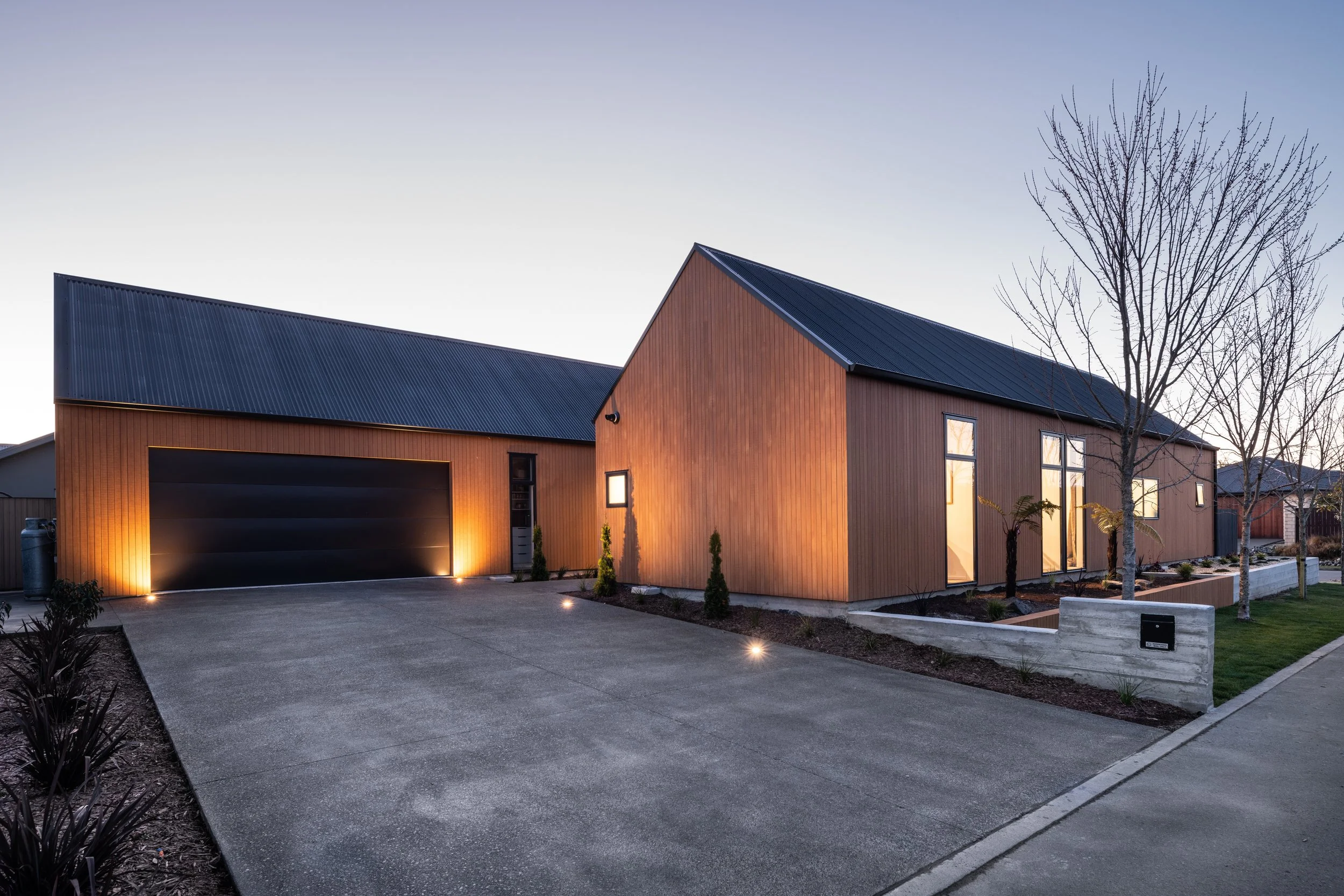 Modern house with wooden exterior and metal roof, front yard with trees and garden beds, driveway leading to garage, and exterior lighting