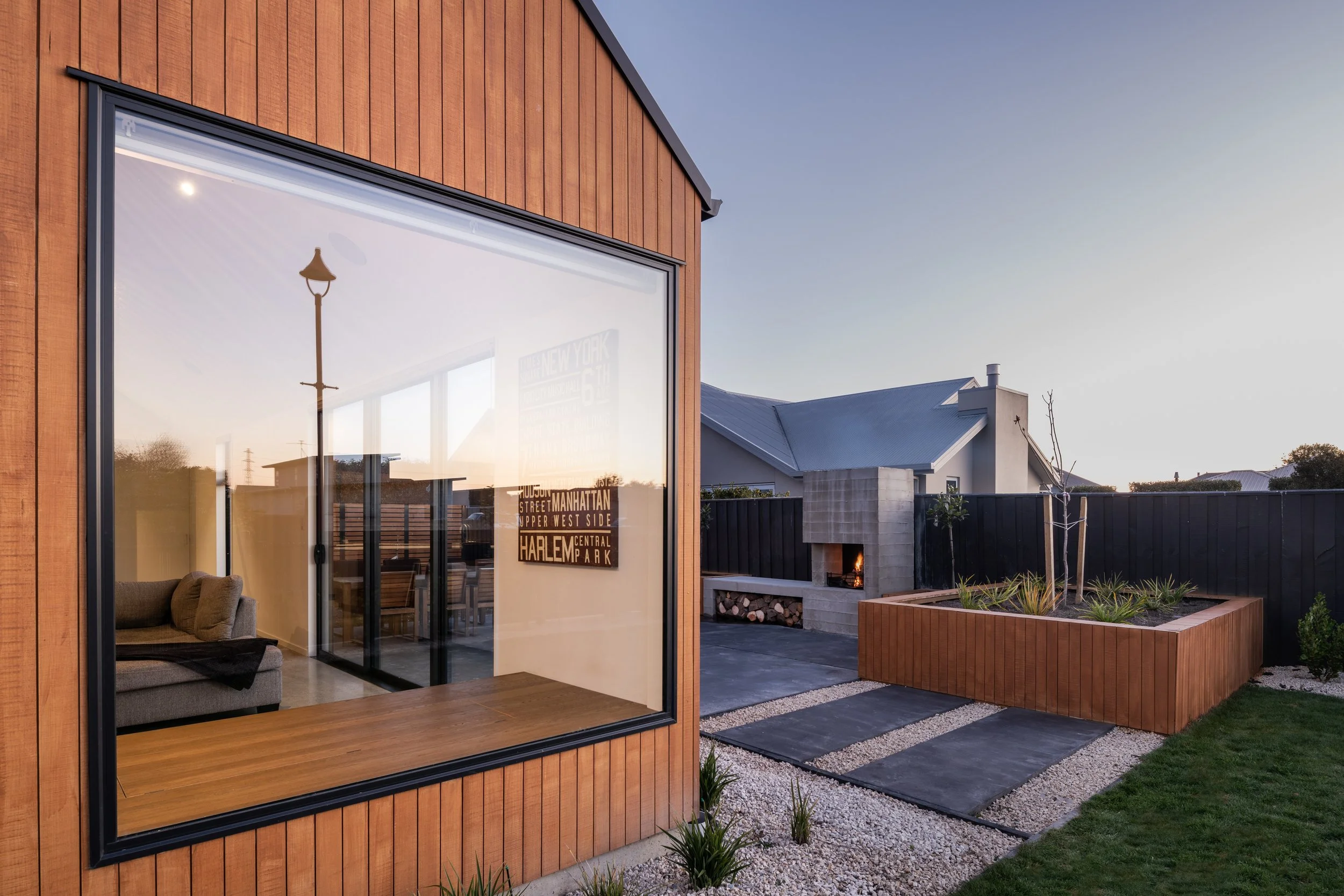 Contemporary backyard with large windowed living space, outdoor fireplace, raised garden bed with small leafless tree, potted plants, and a black fence during sunset.