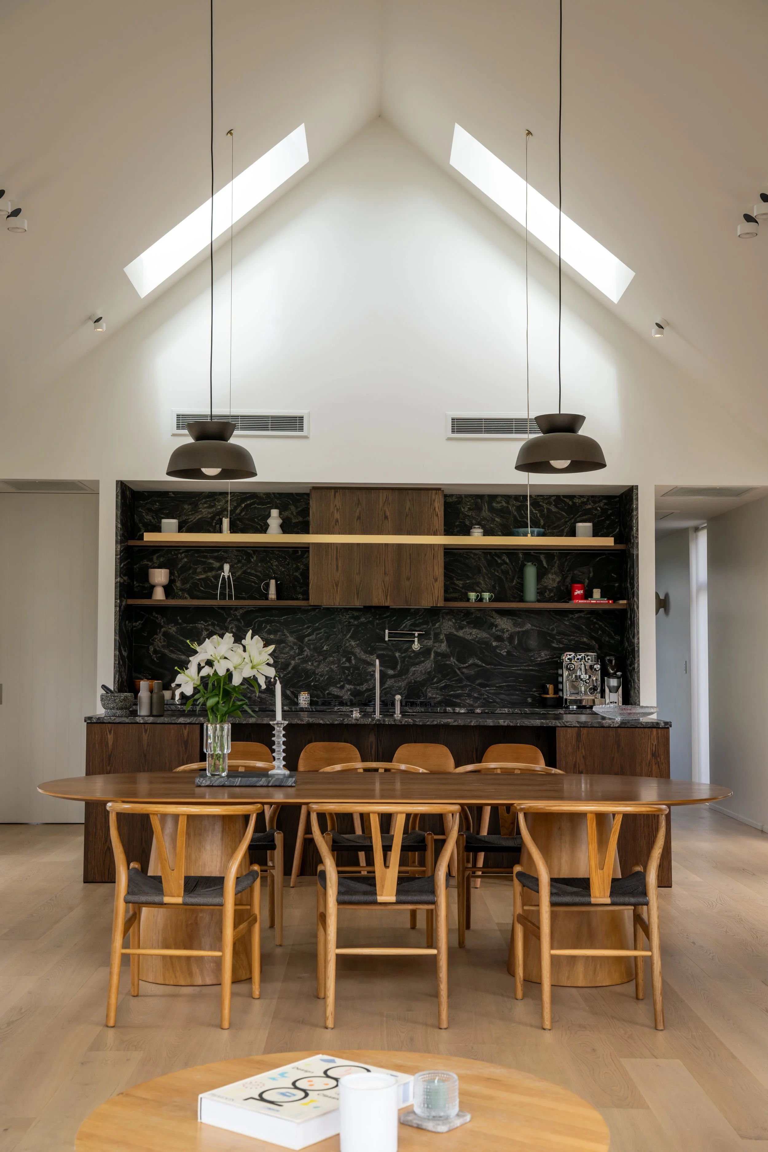 Dining room with wooden table, chairs, black marble kitchen backsplash, and open shelves with decorative items. Skylights in the ceiling, hanging pendant lights, and a flower arrangement on the table.