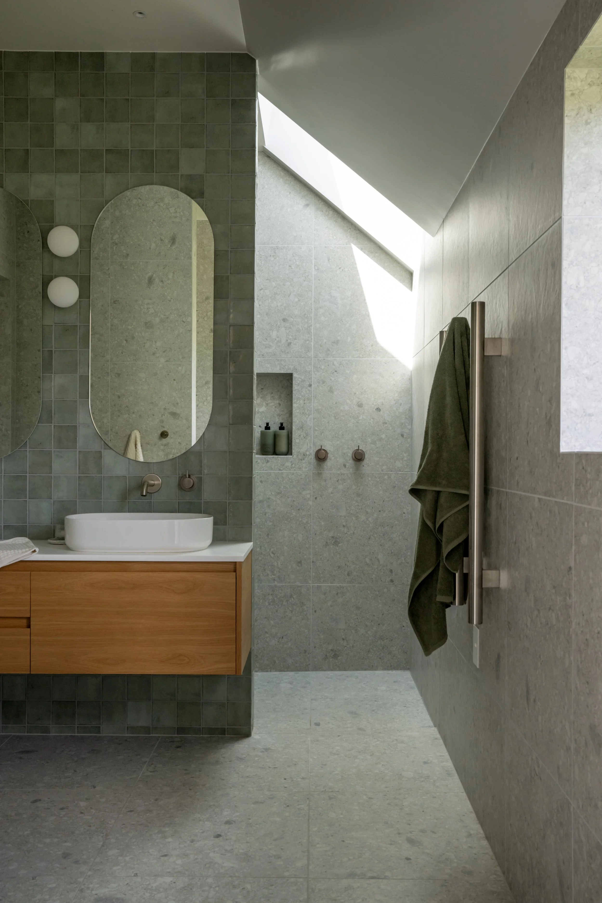 Modern bathroom with light gray tiled walls and floor, wooden vanity with a white vessel sink, large mirror, skylight, and a green towel hanging on a stainless steel towel rack.