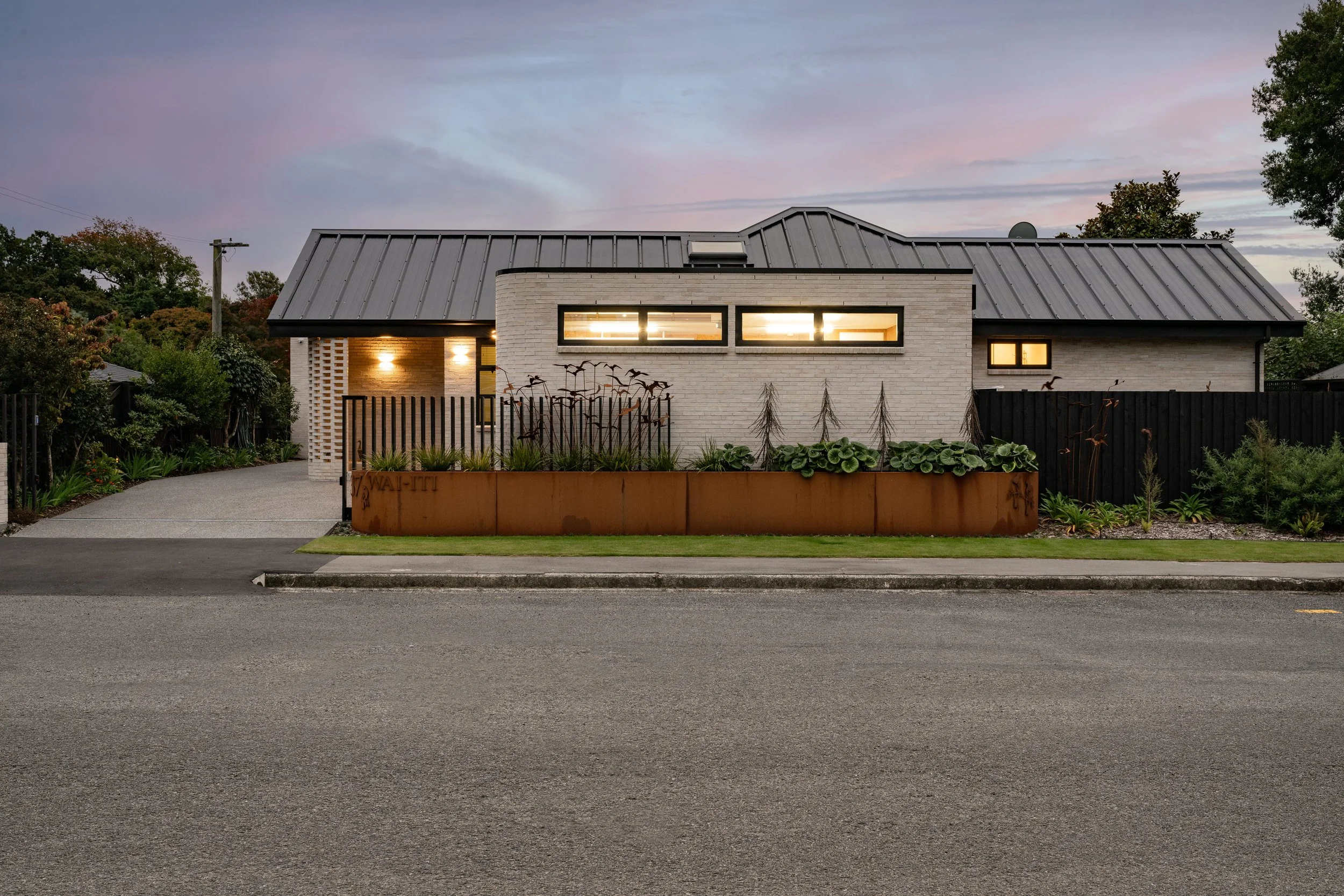 A modern house with a gray metal roof, white brick exterior, and large horizontal windows, at dusk, with front yard and trees in the background.