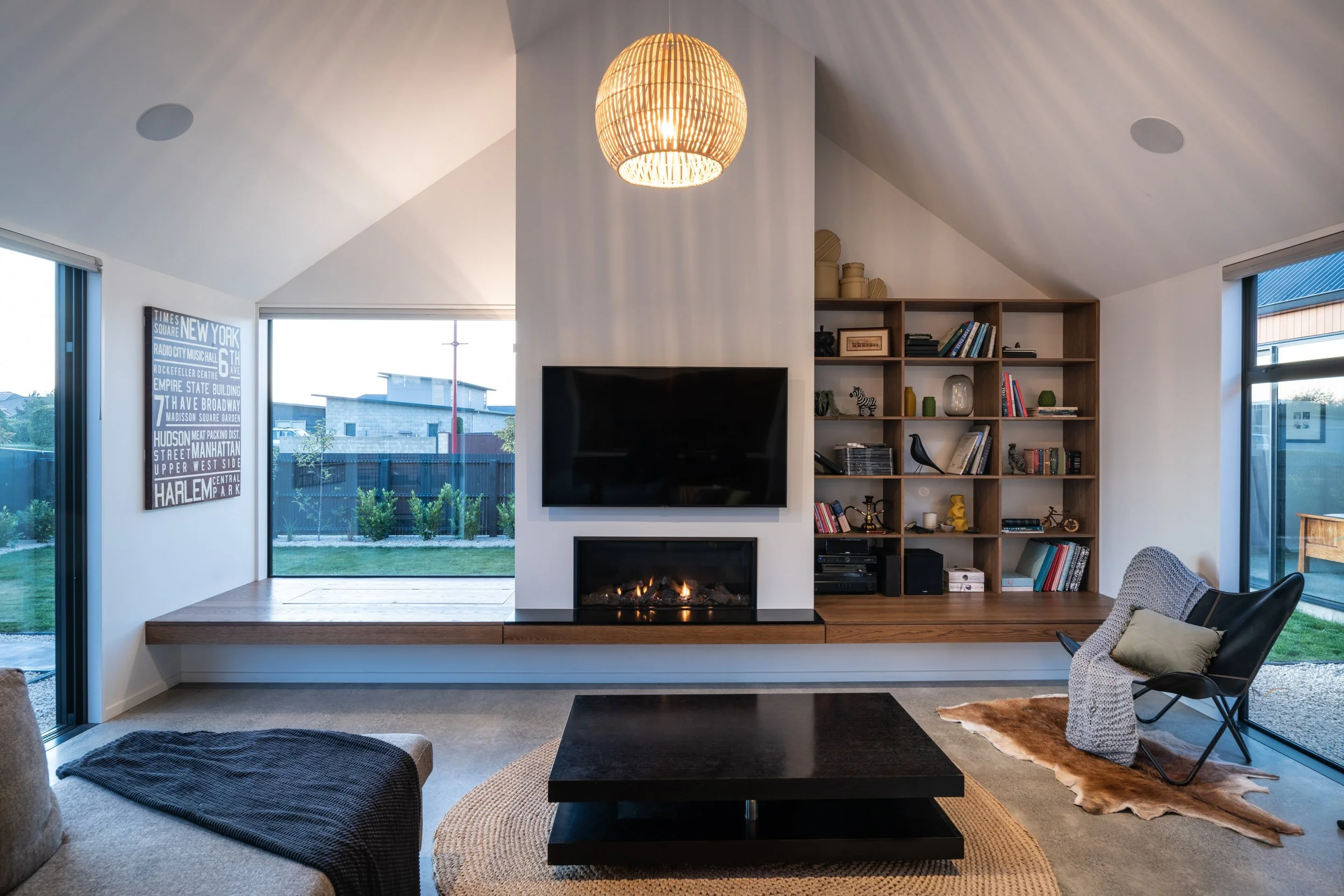 Modern living room with a fireplace, wall-mounted TV, bookshelf, black armchair with a pillow, rug, and large windows showing a backyard.