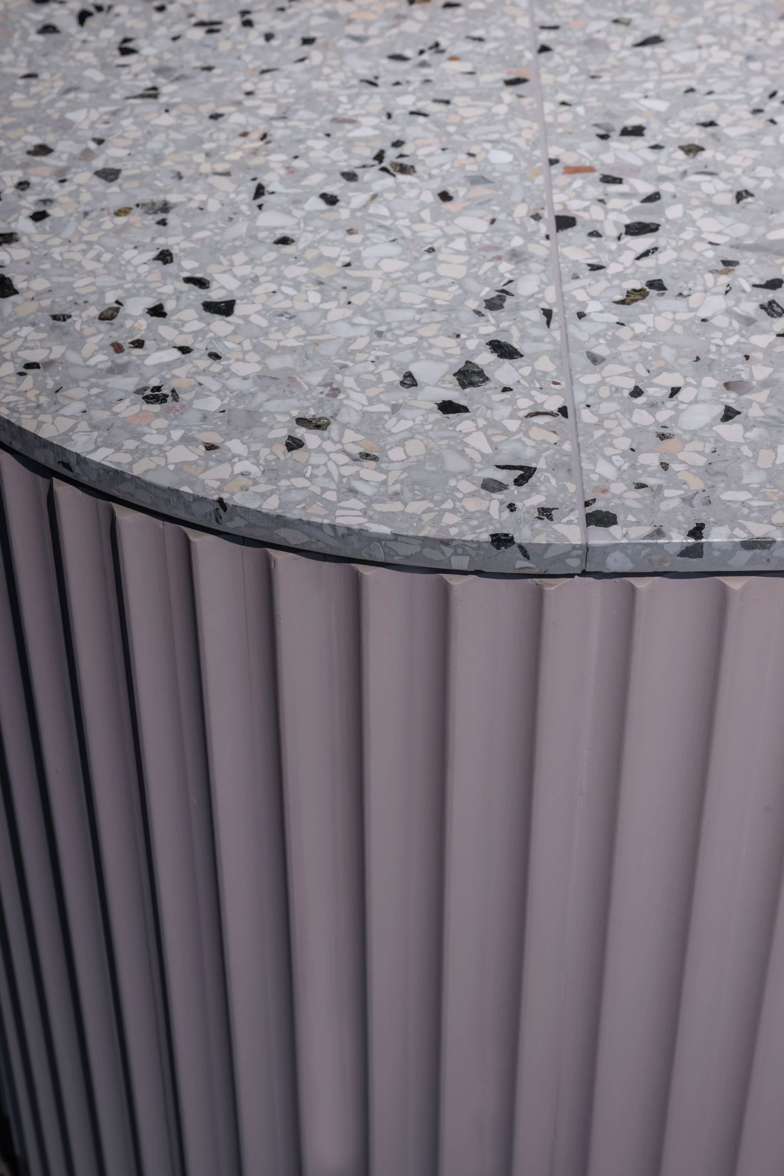 A close-up of a rounded tabletop with a terrazzo surface, featuring small white, black, and gray stone chips, above vertical, ribbed pinkish-beige paneling.