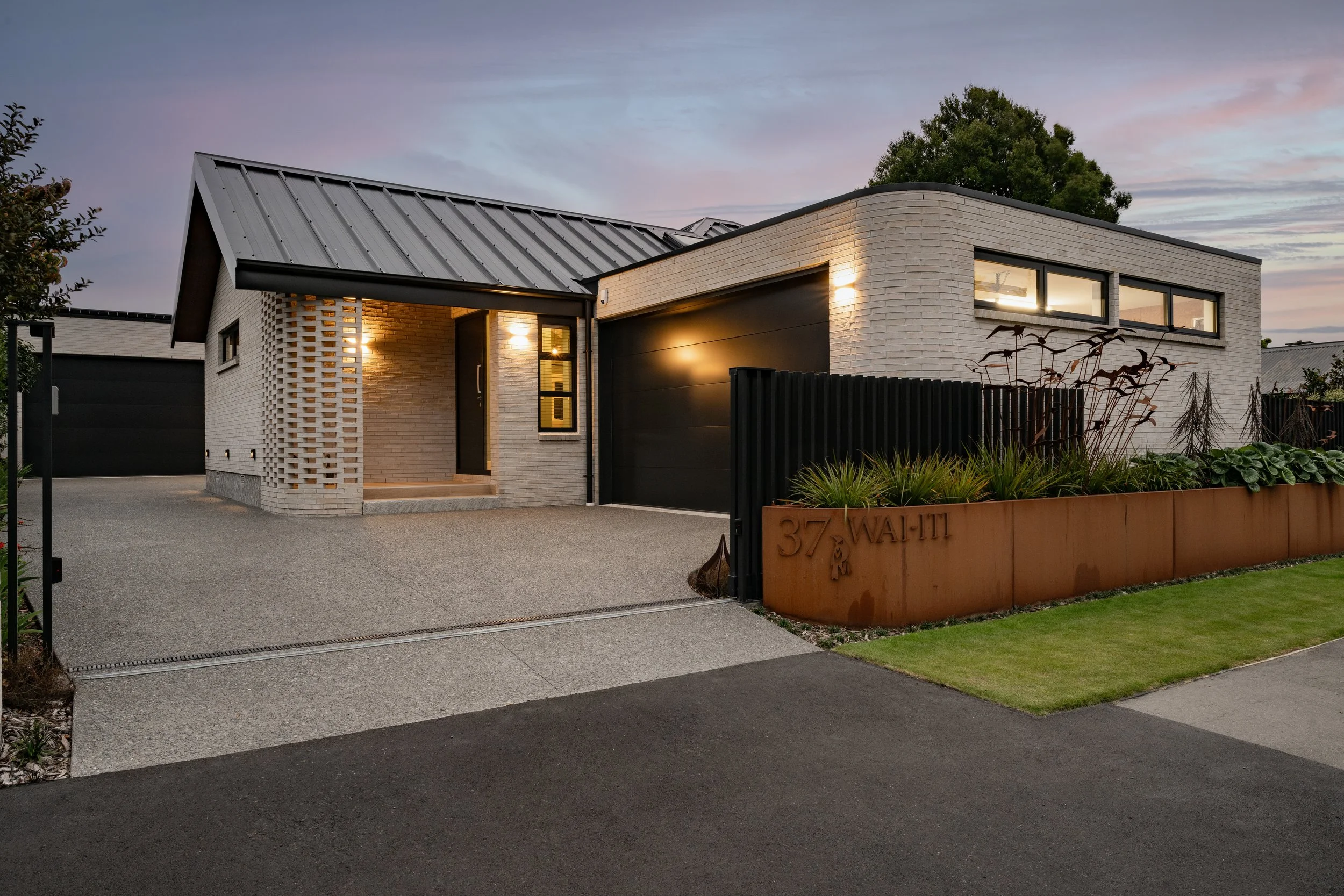 Modern house with beige brick walls, black accents, and large windows, illuminated by exterior lighting at dusk, surrounded by a garden and a concrete driveway.