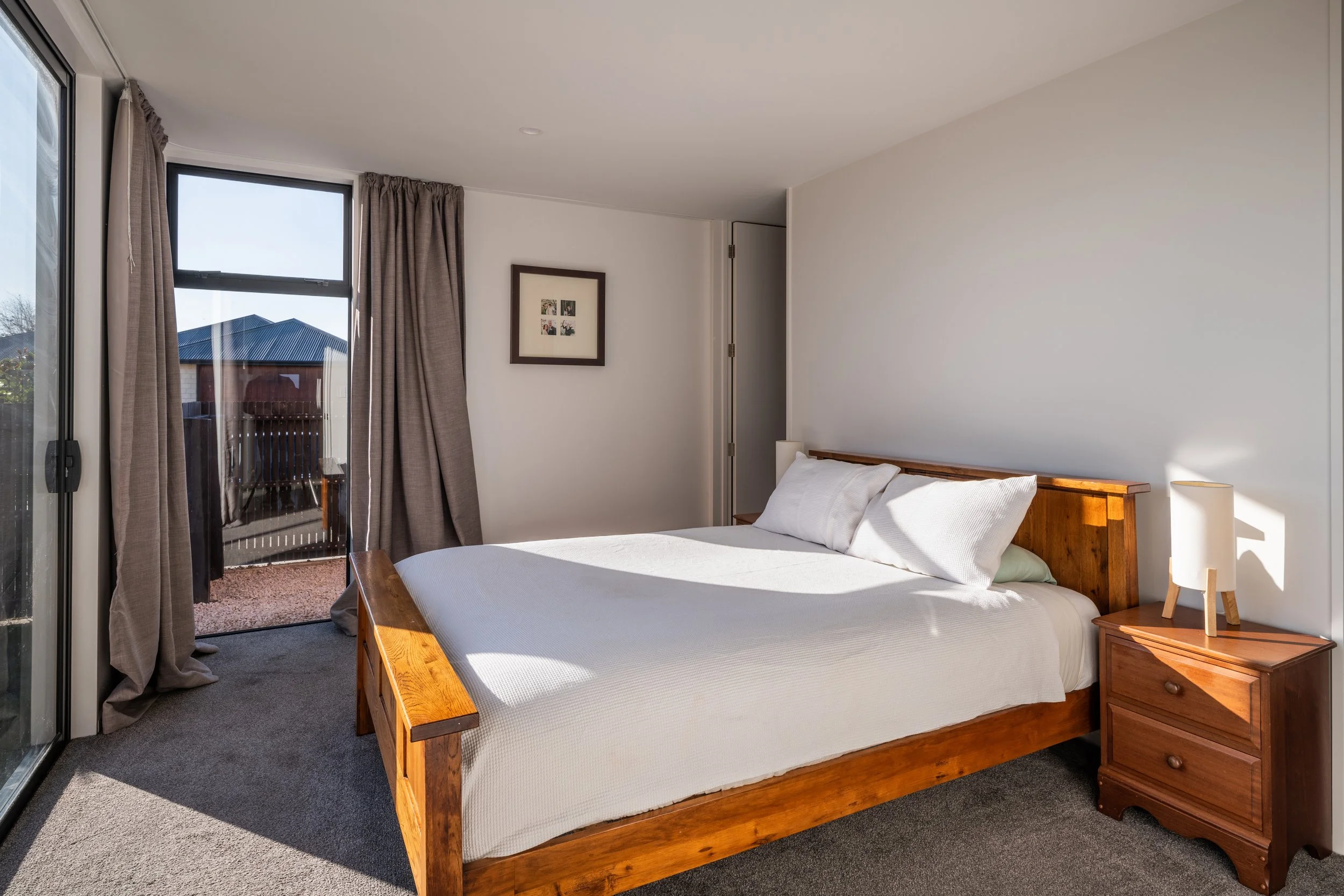 A neatly made bed with white bedding and pillows sits in a bedroom with a wooden headboard and nightstand. Sunlight streams through large glass doors with brown curtains, leading to a small balcony outside. A framed picture hangs on the white wall to