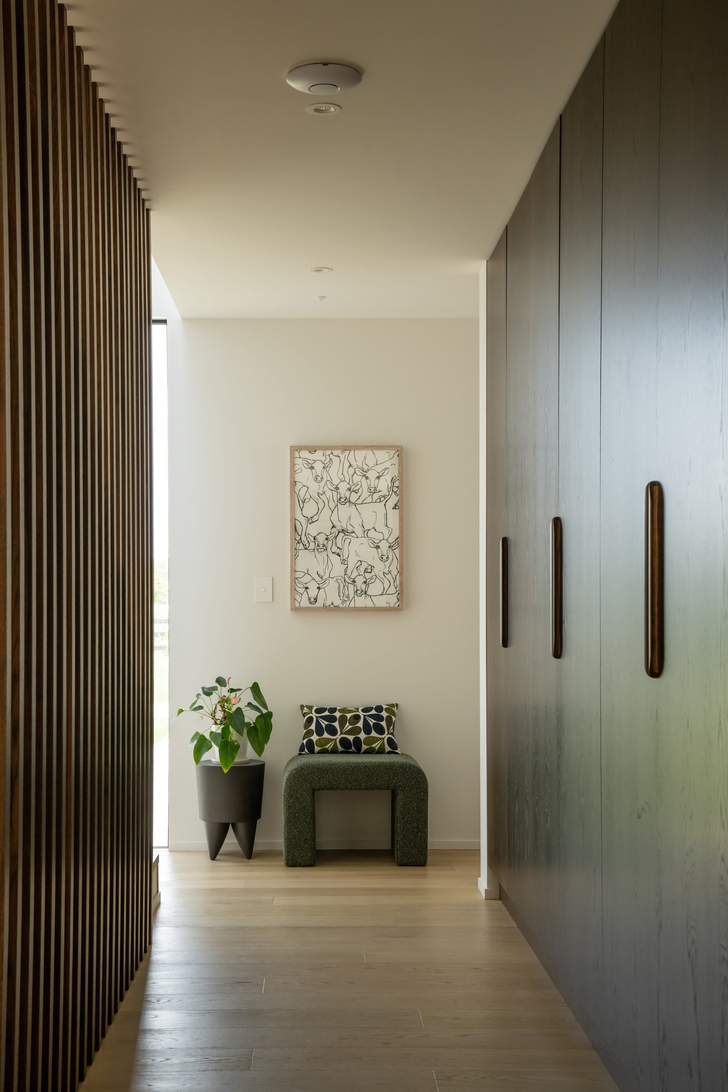 Modern entry hallway with light wood flooring, wooden slat wall on the left, a dark wood closet on the right, a small green seat with textured fabric, a potted plant on a gray pedestal, and framed artwork of deer sketches on the back wall.