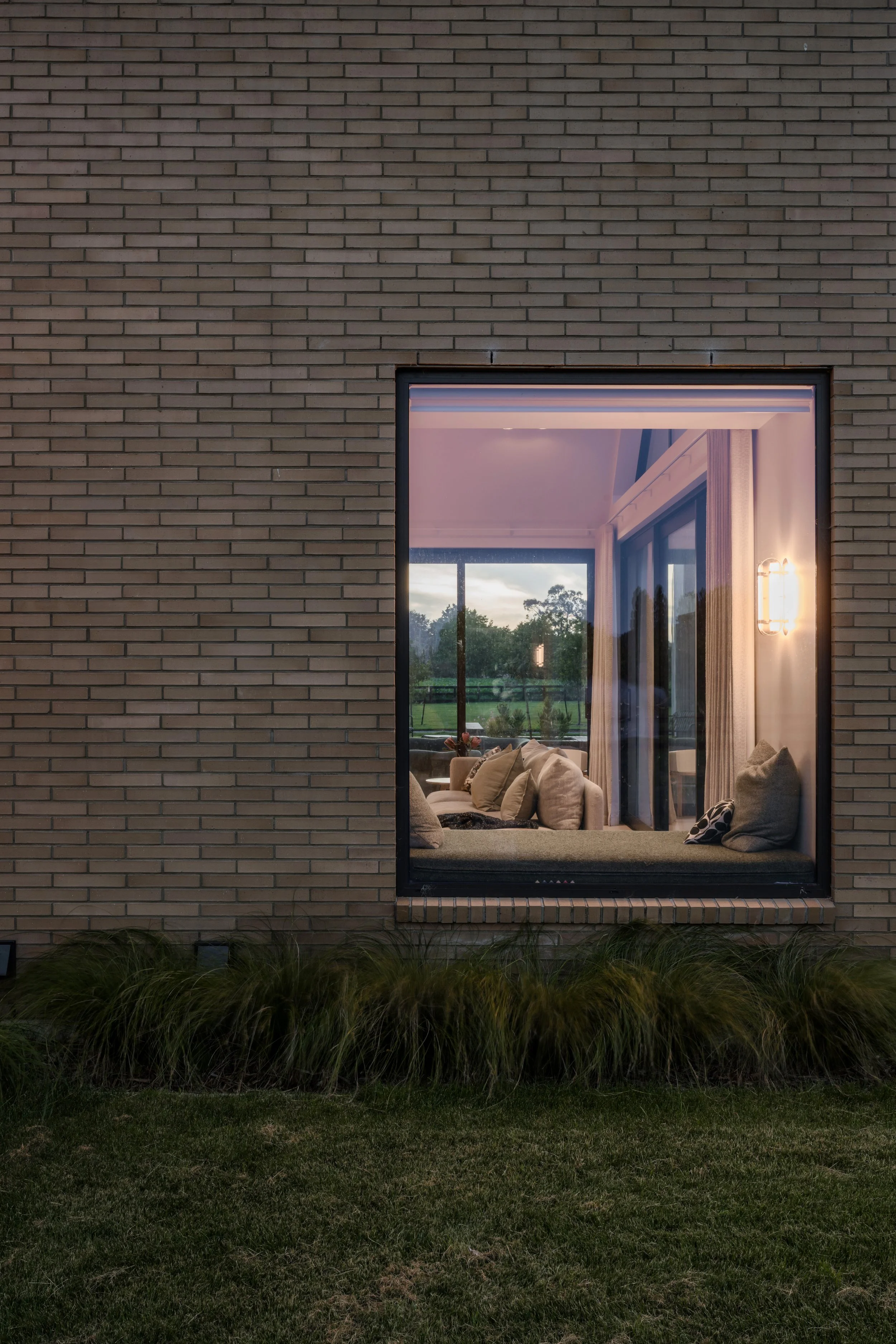 A view of a brick house exterior with a large window showing a living room with a sofa and pillows, illuminated by a wall-mounted light. Outside the window, a green landscape with trees and a cloudy sky is visible during dusk or early evening.