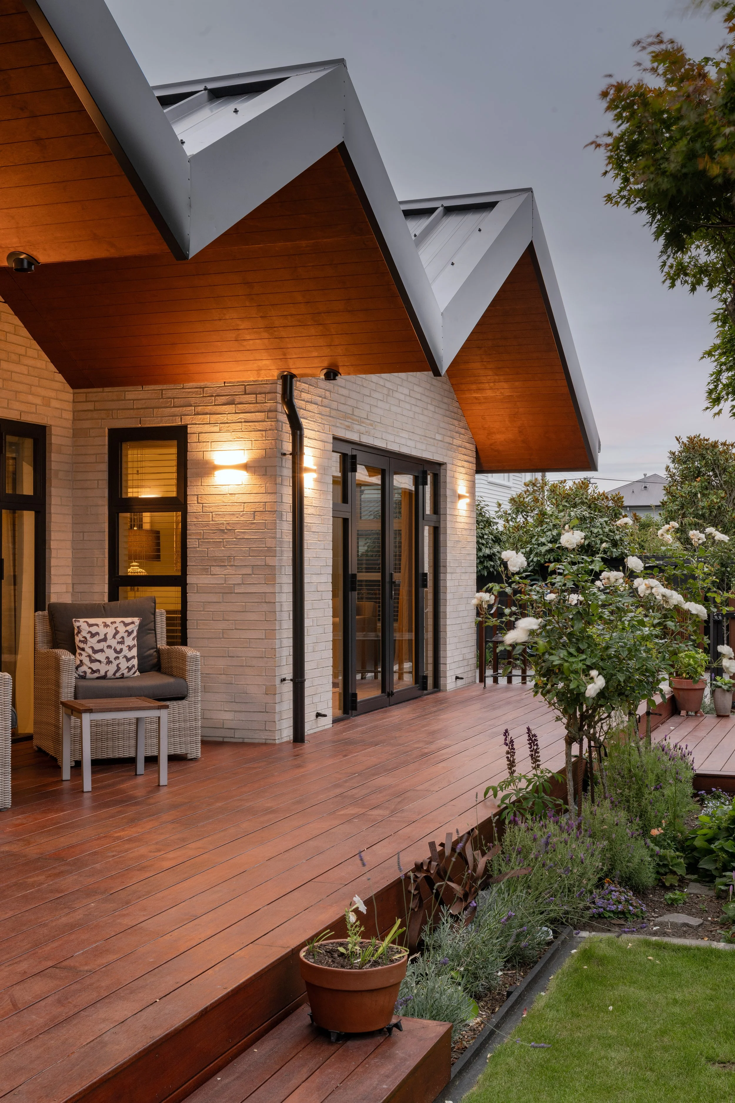 A house with a wooden deck, outdoor seating, and a garden with flowering plants and bushes, under an overcast sky.