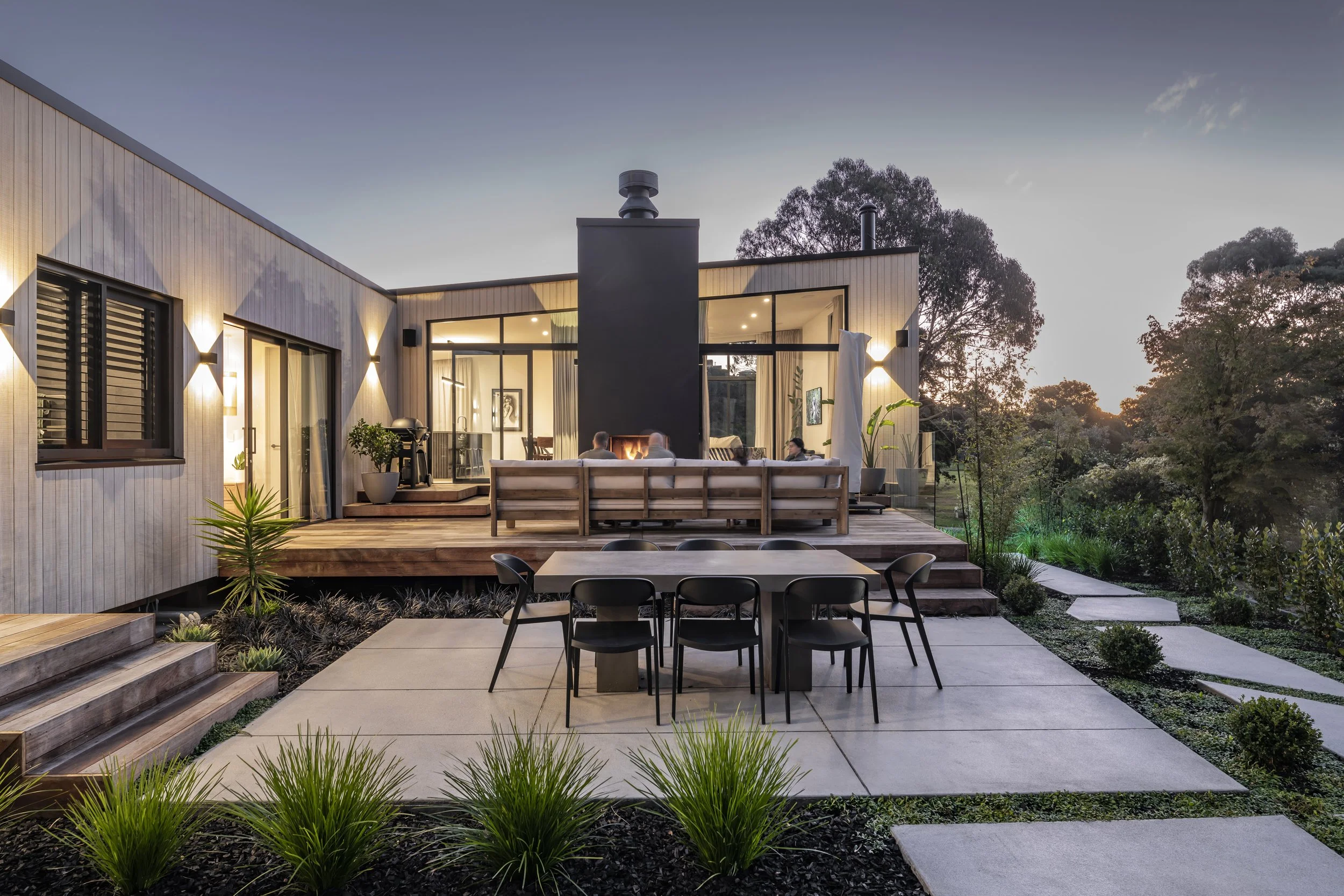 Modern house with outdoor patio, dining table, and seating area with fireplace, surrounded by lush greenery at dusk.