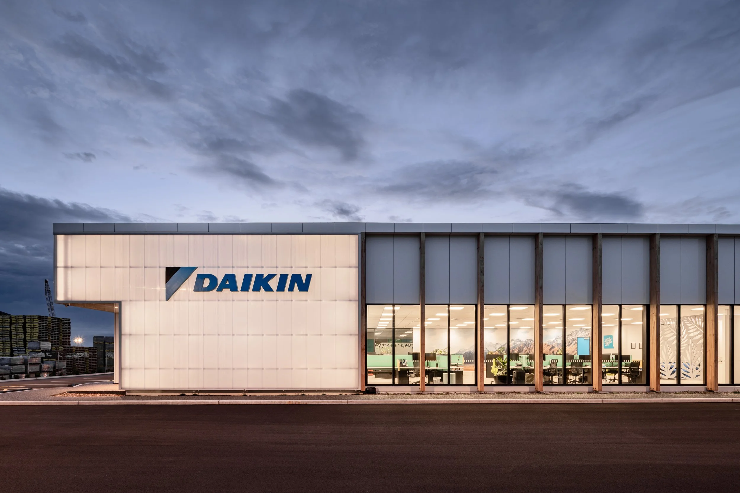 Exterior of a modern building with large windows, displaying an illuminated interior showroom for Daikin, a heating and cooling company. The sky above is partly cloudy at dusk.