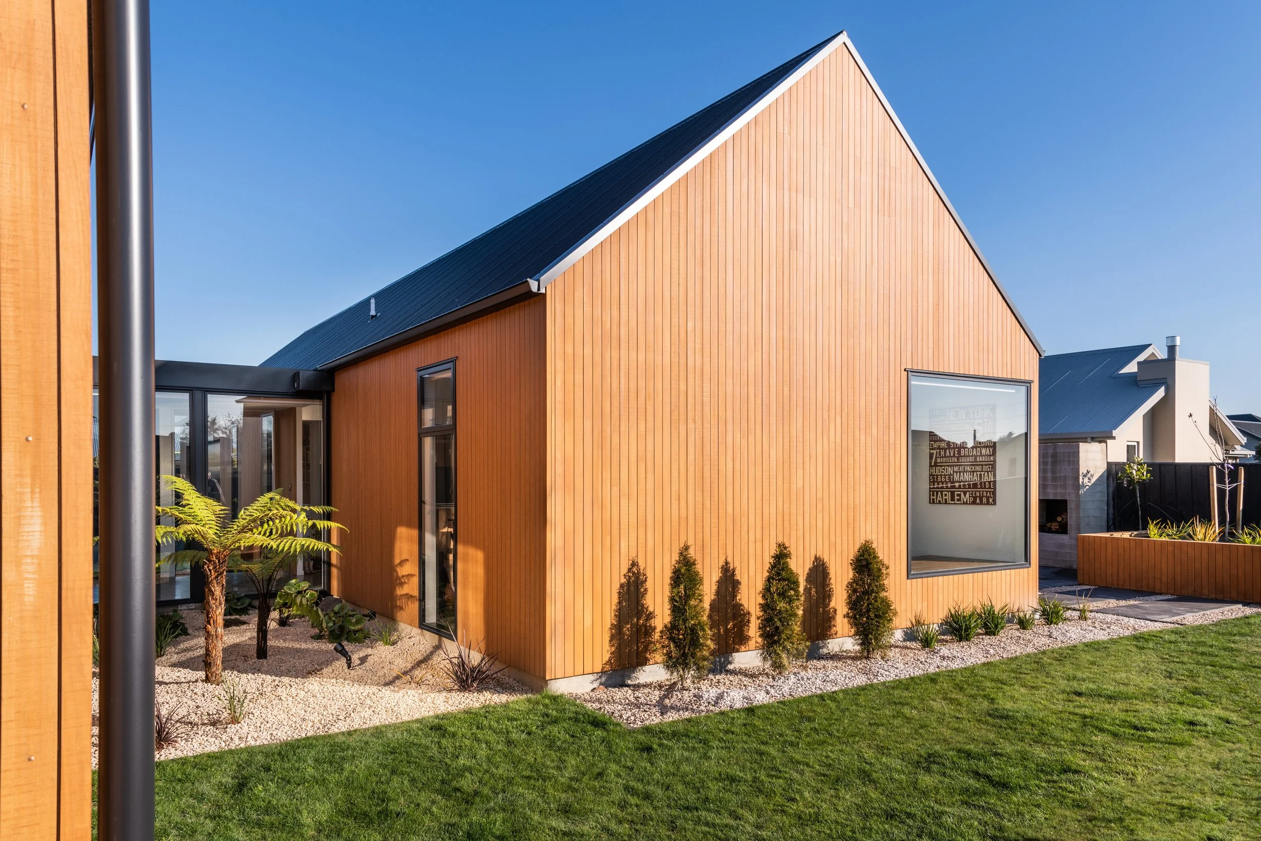 Modern house with wooden exterior walls, metal roof, large windows, landscaped yard with small plants and trees, under clear blue sky.