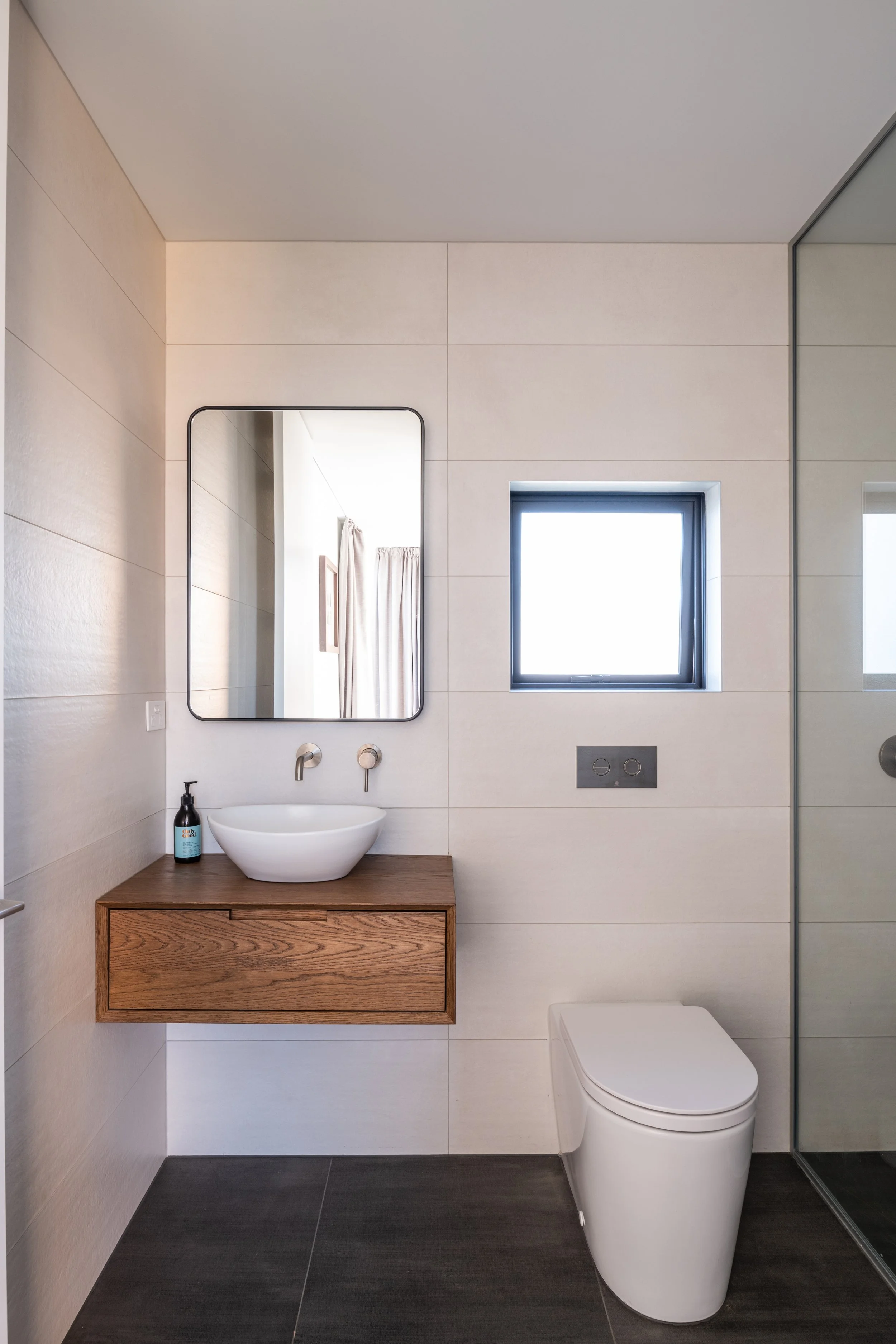 Modern bathroom with a wooden vanity, white vessel sink, rectangular mirror, small window, and a white toilet.