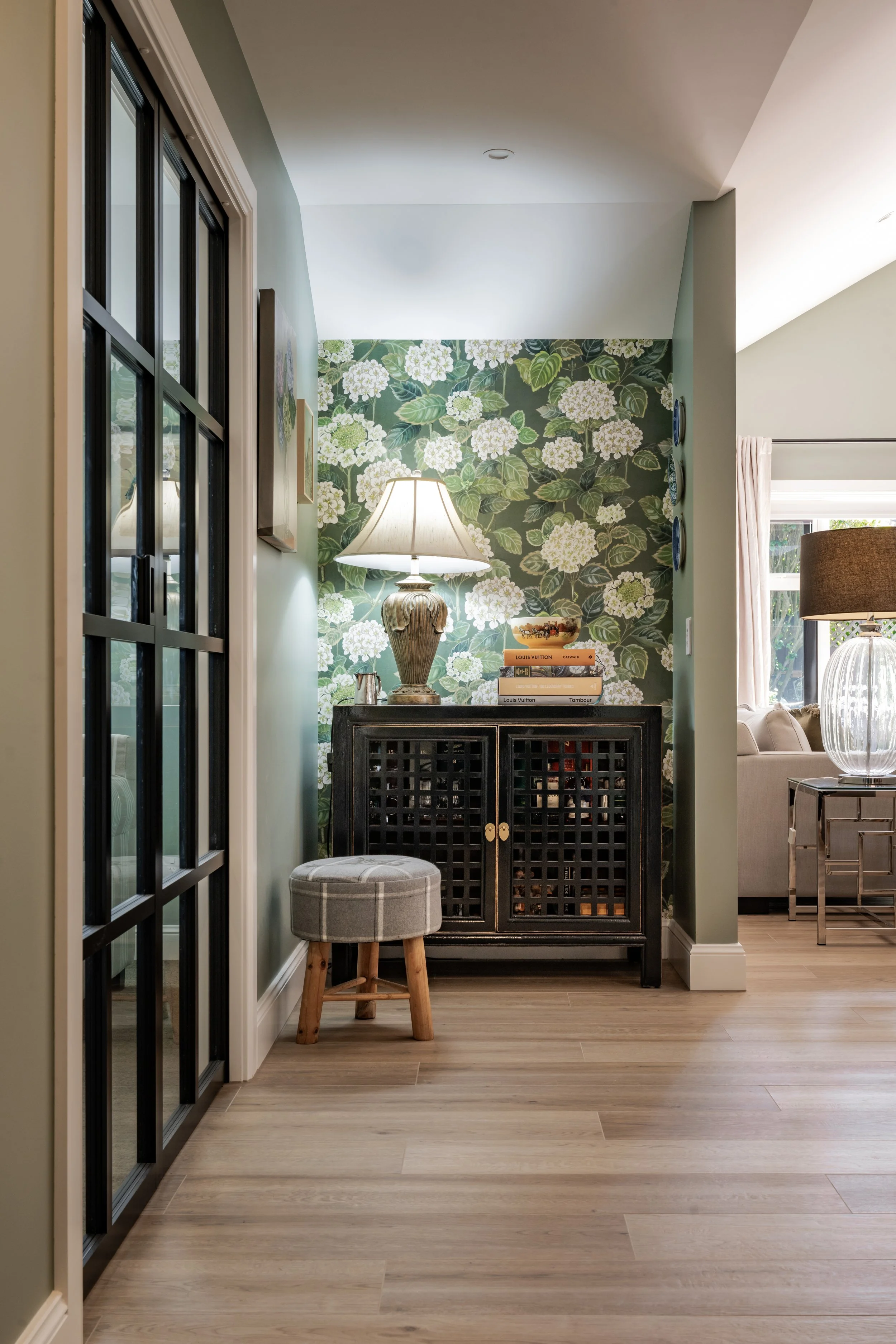 A cozy corner with a dark wooden cabinet, a table lamp, a small plaid cushioned stool, and artwork on the green floral wallpapered wall in a well-lit home interior.