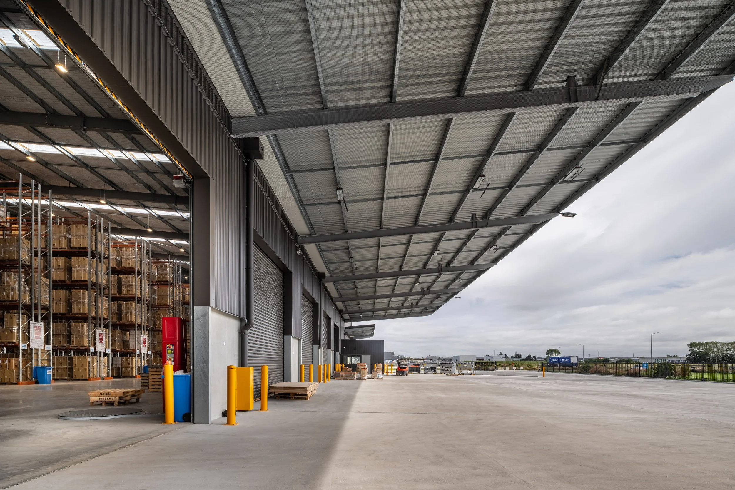 Exterior view of a warehouse with open loading dock doors, metal roof, and storage shelves filled with boxes inside. There is a wide paved area outside with some trucks in the background.