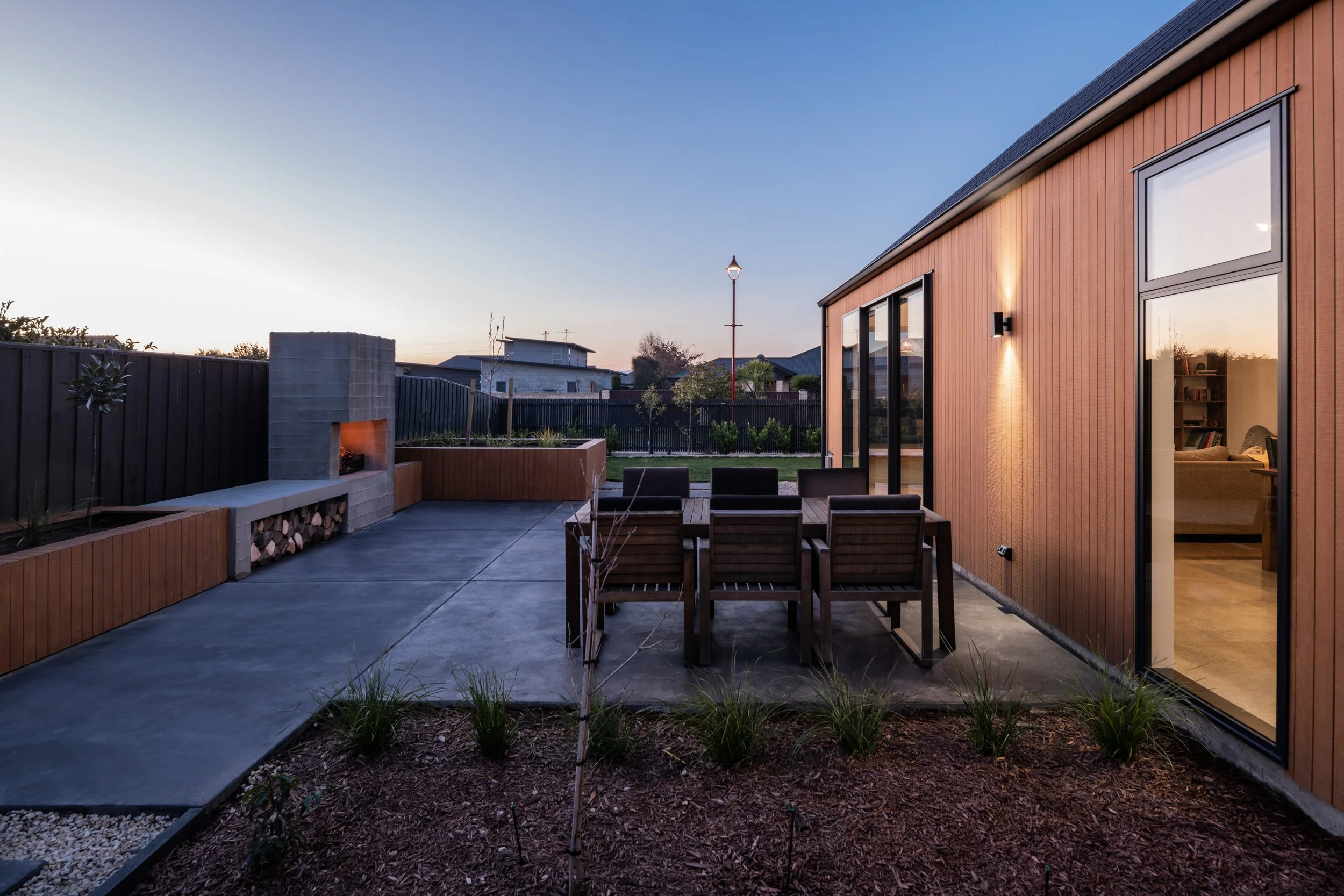 Backyard patio with outdoor fireplace, wooden furniture, garden bed, and view of neighboring houses at dusk.