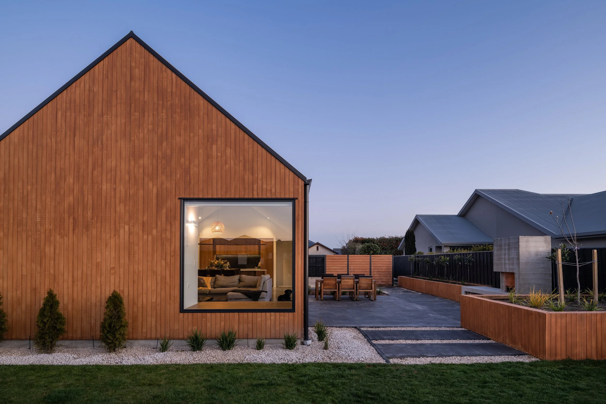 Modern house with wooden exterior, large window showing living room inside, outdoor dining area, landscaped yard with small trees and planters, and neighboring houses in the background during early evening.