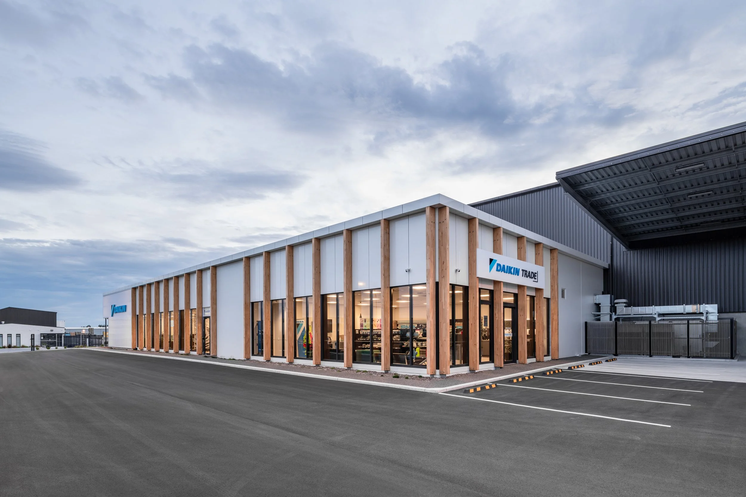 Modern commercial building with wooden and white exterior, glass windows, and a sign that reads 'Daikin Trade', with a parking lot in front and cloudy sky above.