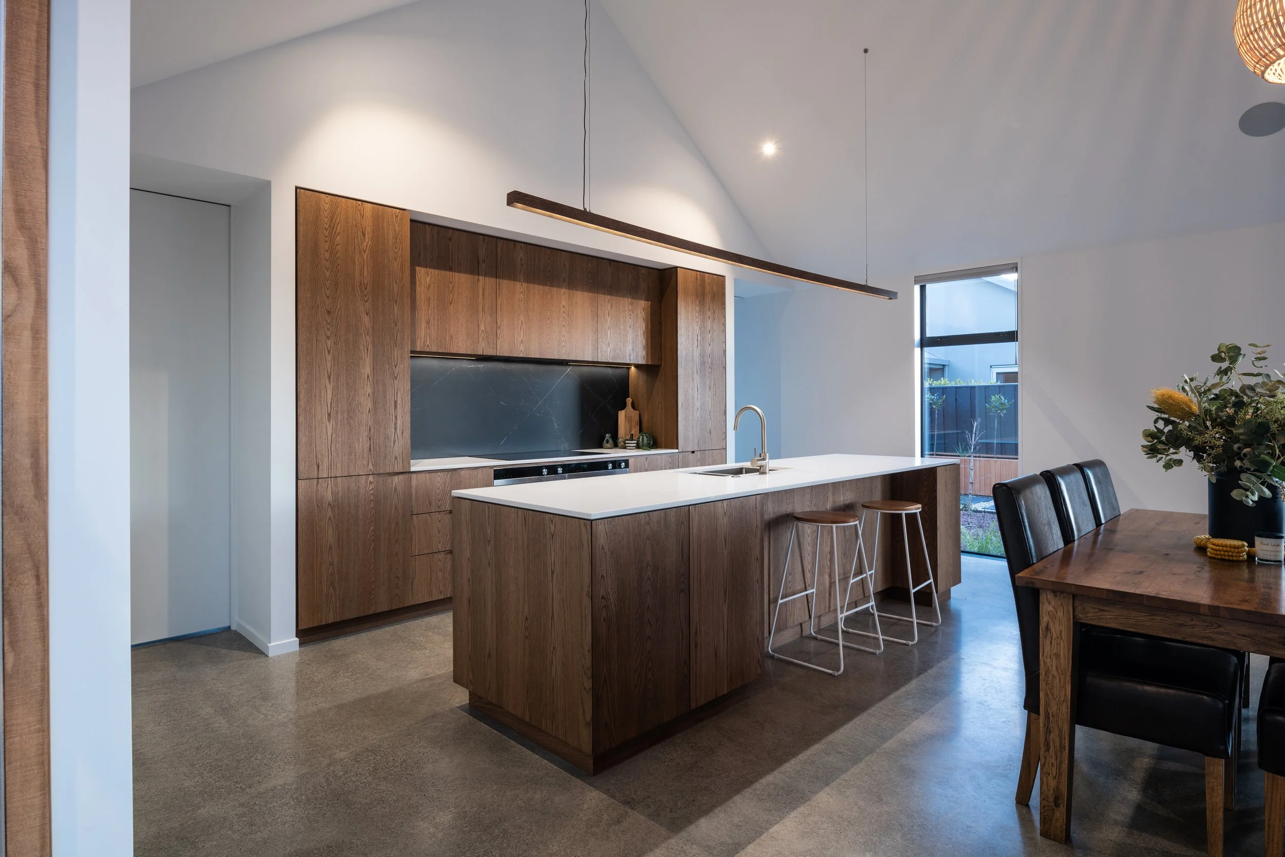Modern kitchen with wooden cabinetry, white island with sink, black backsplash, and a wooden dining table with black chairs, well-lit with natural light.
