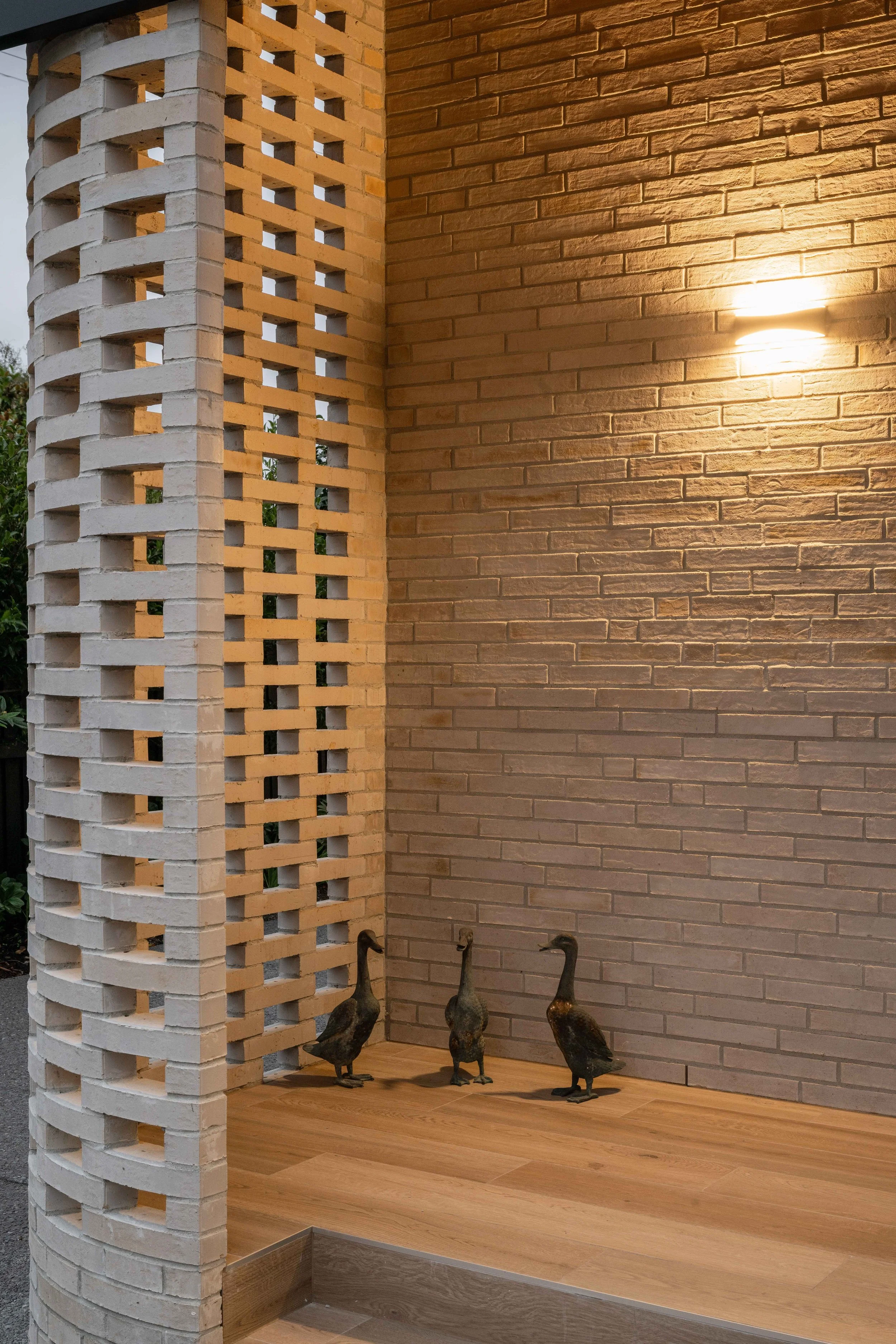 A corner of a room featuring a white brick column and a brown brick wall illuminated by a wall-mounted light, with three duck statues on the wooden floor.