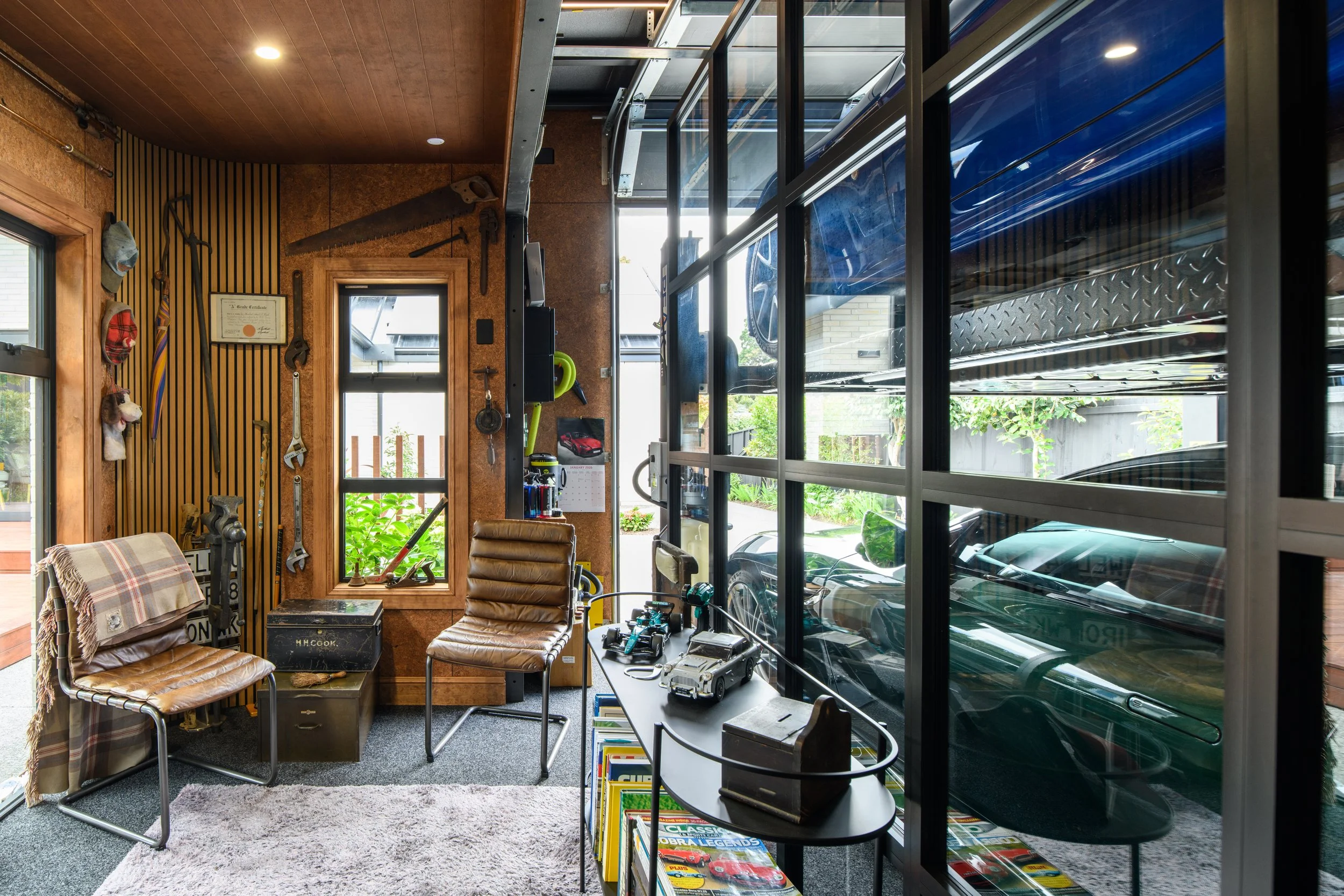 Garage workshop with tools on the wall, a bookshelf with car magazines, and vintage cars visible through large windows.