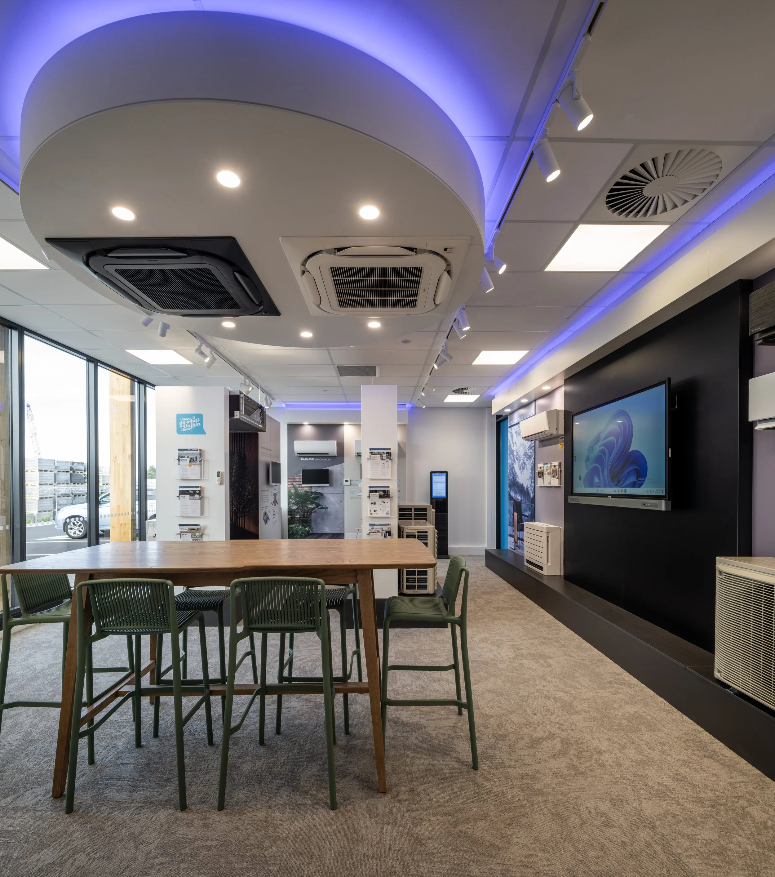 Modern office or conference room with a wooden table, green chairs, large screens, and ceiling air conditioning units.