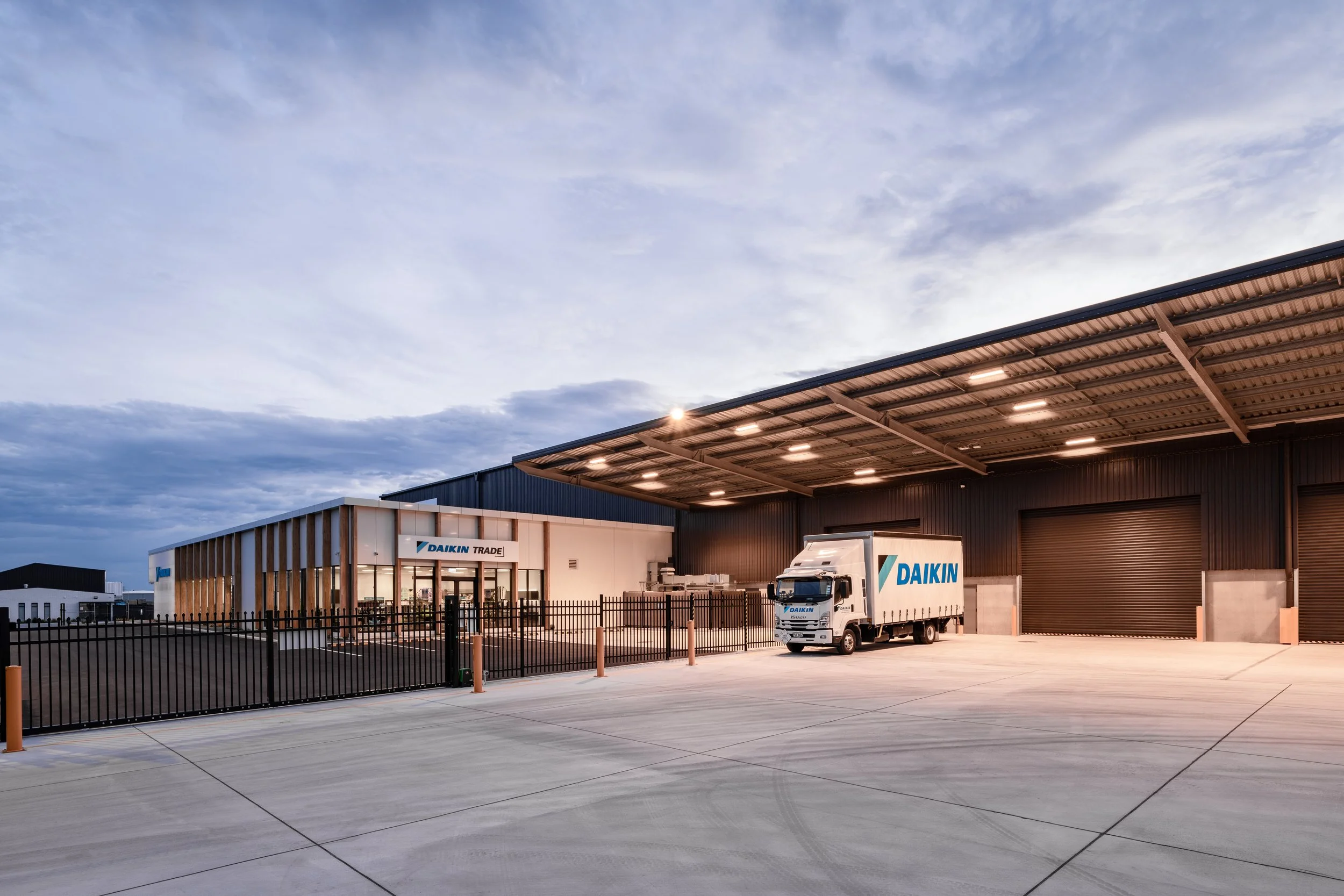 Outdoor view of a modern commercial building with a truck parked in front, illuminated by overhead lights during dusk or early evening, with a sky filled with clouds.