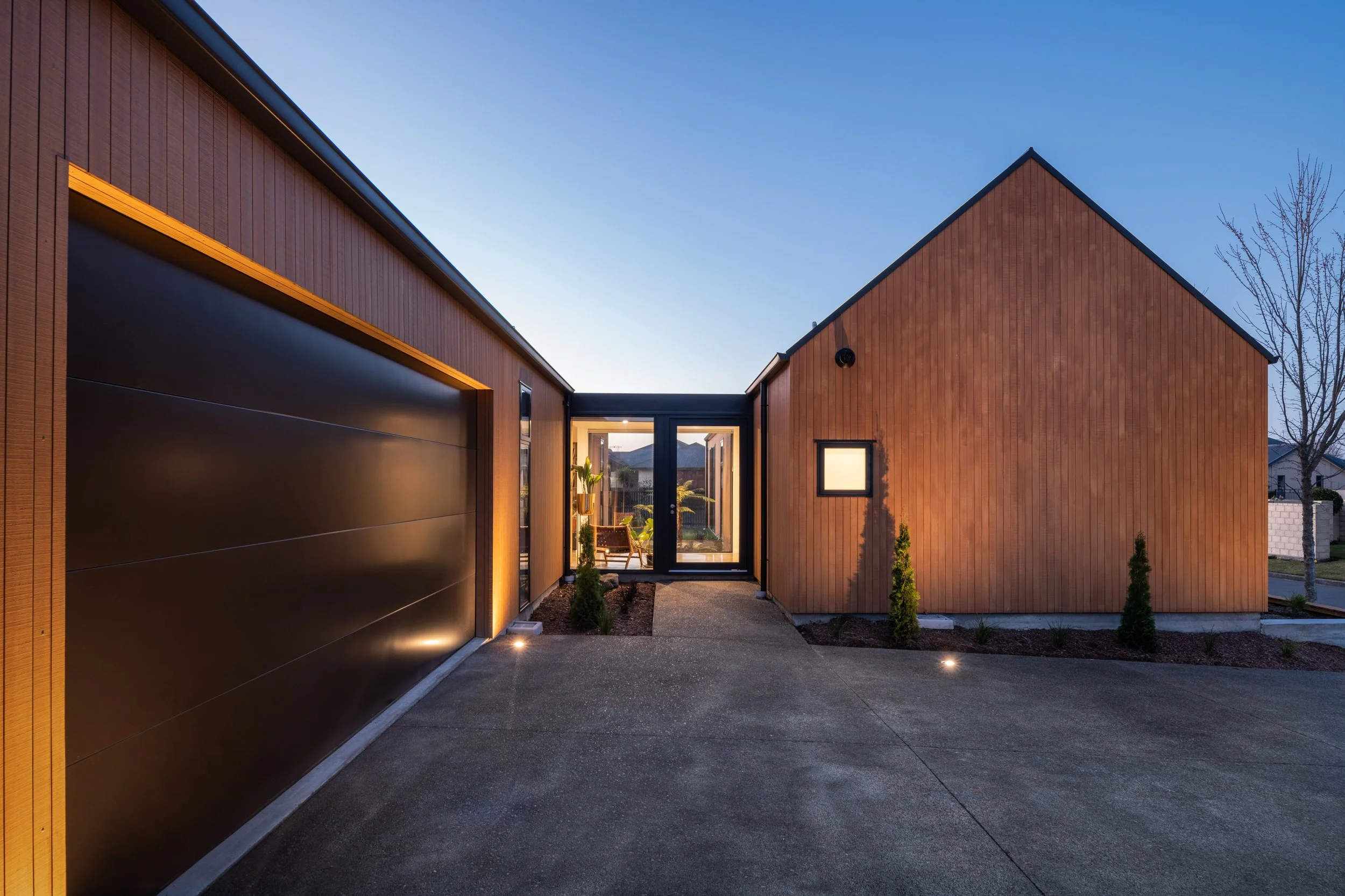 Modern house exterior at dusk with wooden siding, black garage door, and glass front door illuminated from inside.