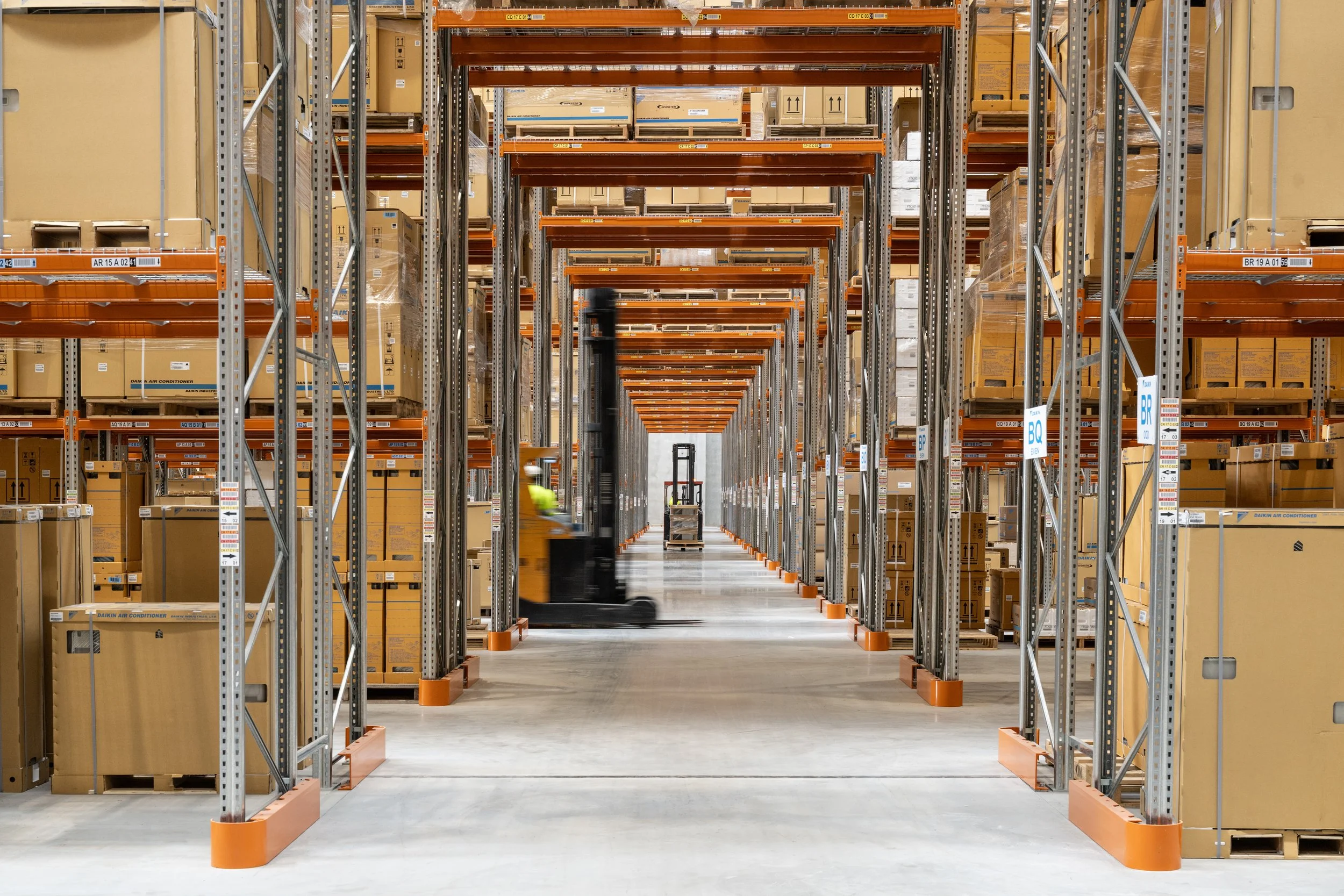 View of warehouse aisles with tall metal shelving filled with boxed products, a person operating a forklift in the aisle.