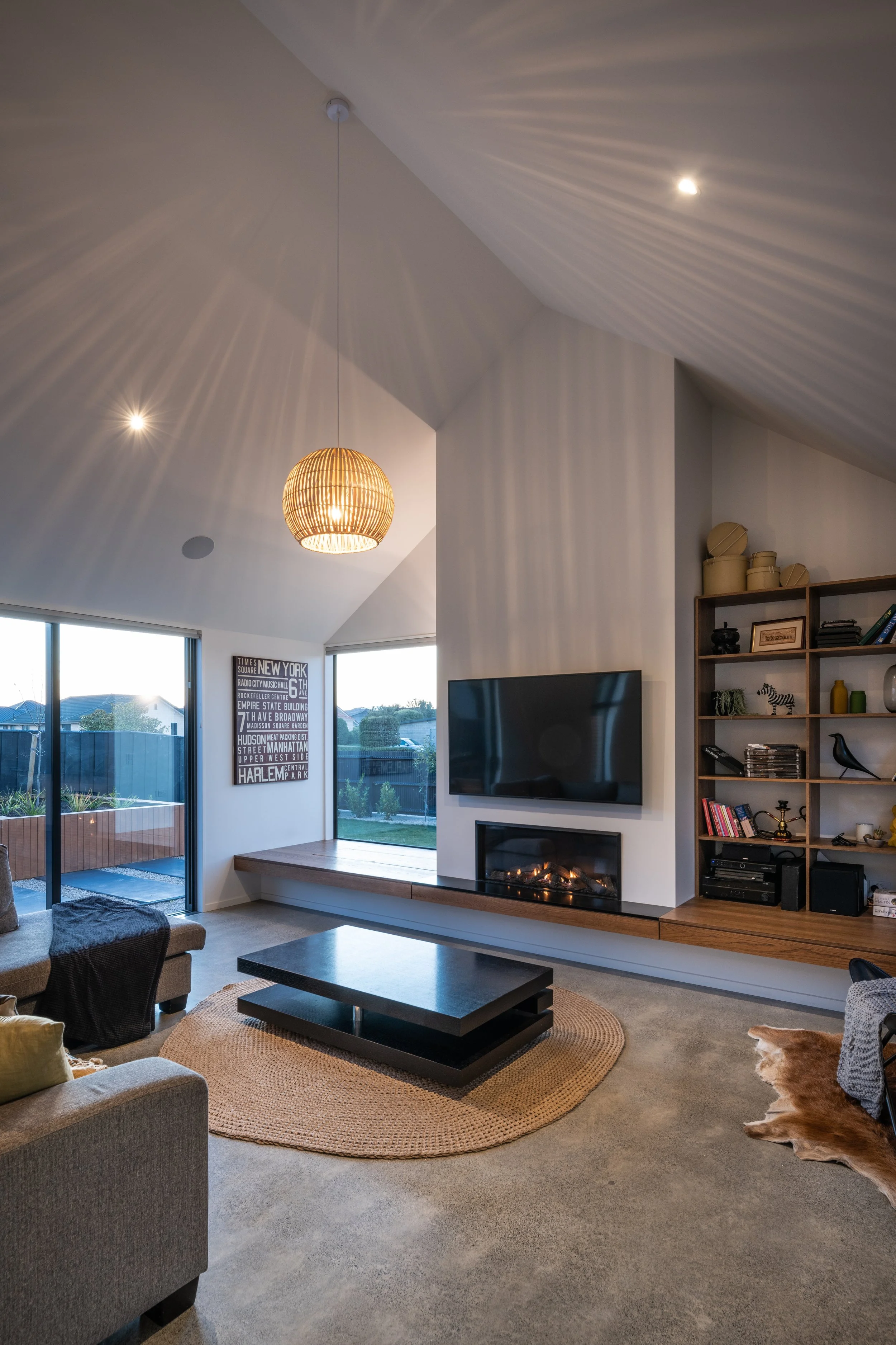 Modern living room with vaulted ceiling, a hanging woven light, a mounted flat-screen TV above a fireplace, a large window with city view, a bookshelf with decor, a black coffee table on a round rug, and a beige couch with pillows.