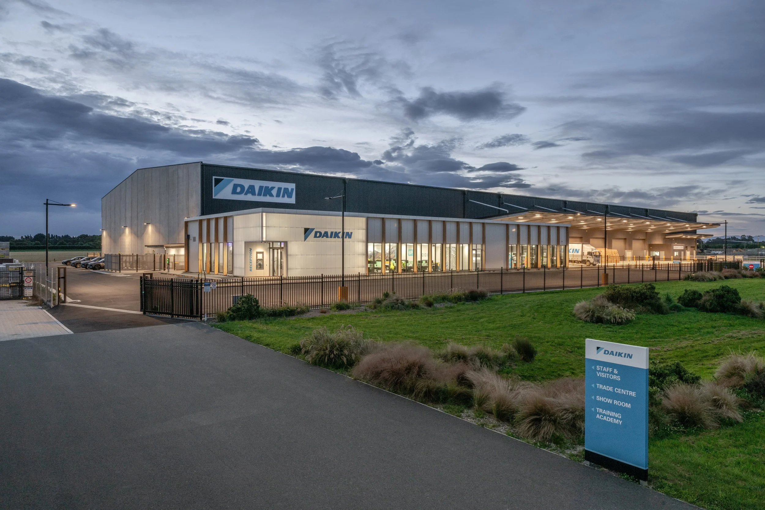 Exterior view of a large commercial building with a sign reading 'Daikin', illuminated under cloudy evening sky, surrounded by a parking lot, fencing, and landscaped bushes.